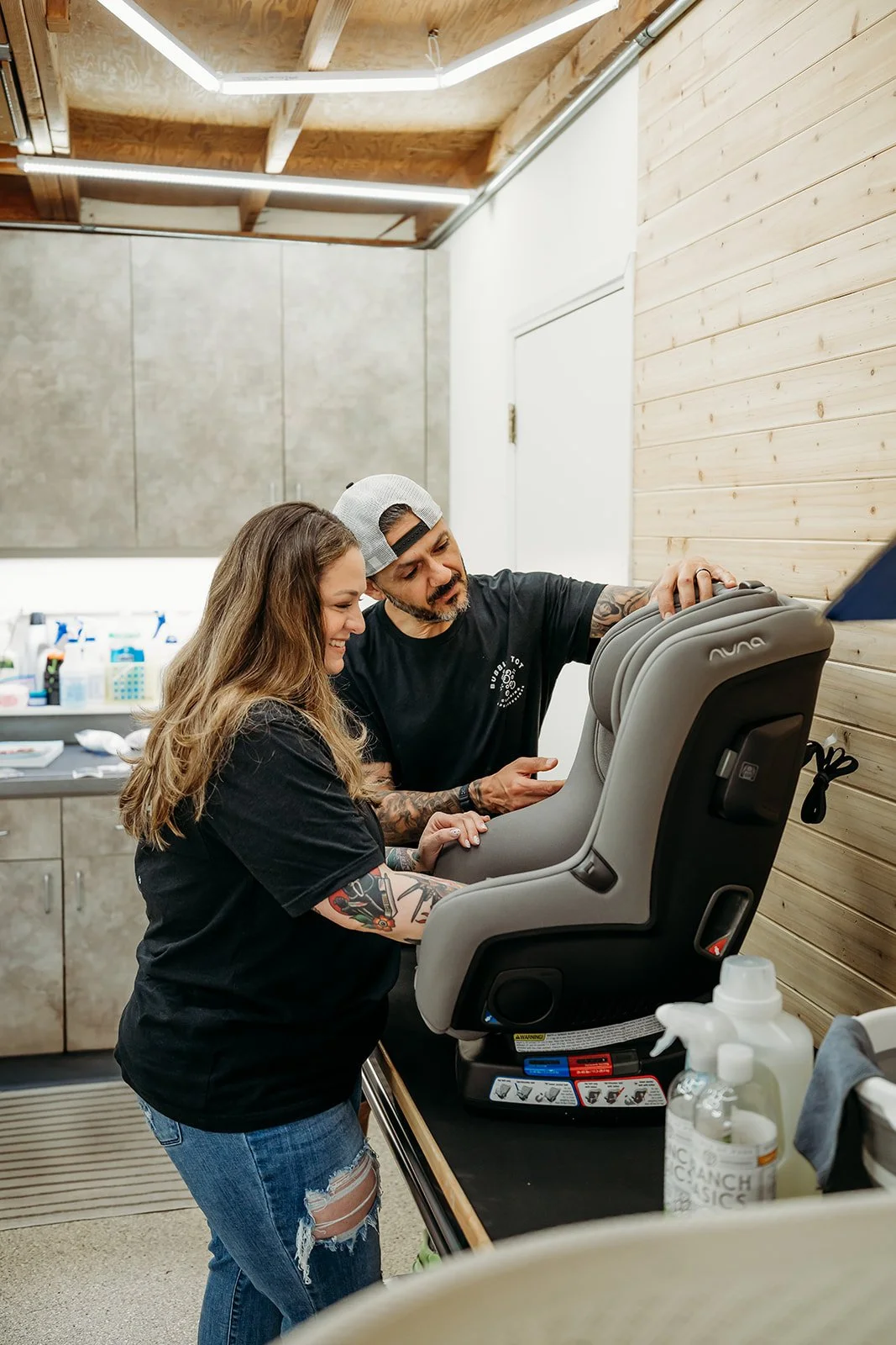 A male and female working together inspecting a child's car seat in a room with wood-paneled walls and cleaning supplies on the counter.
