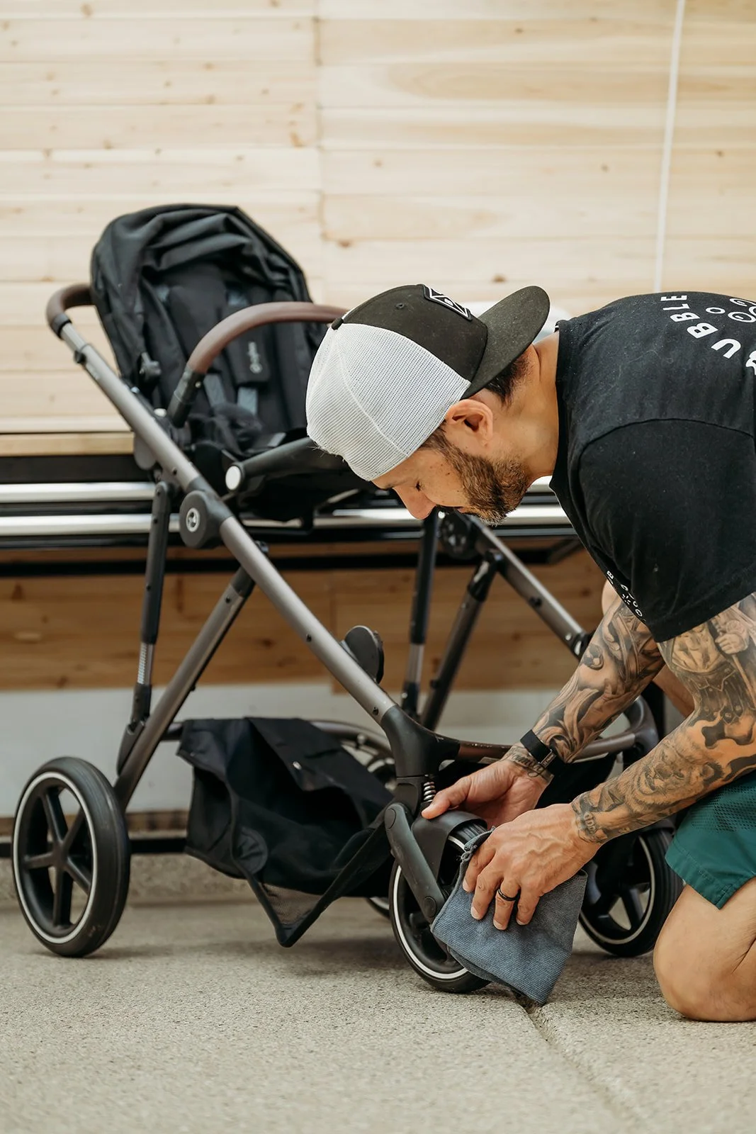 A man with tattoos, wearing a black cap and black T-shirt, is cleaning the floor with a cloth in front of a black stroller, in a room with wooden walls.