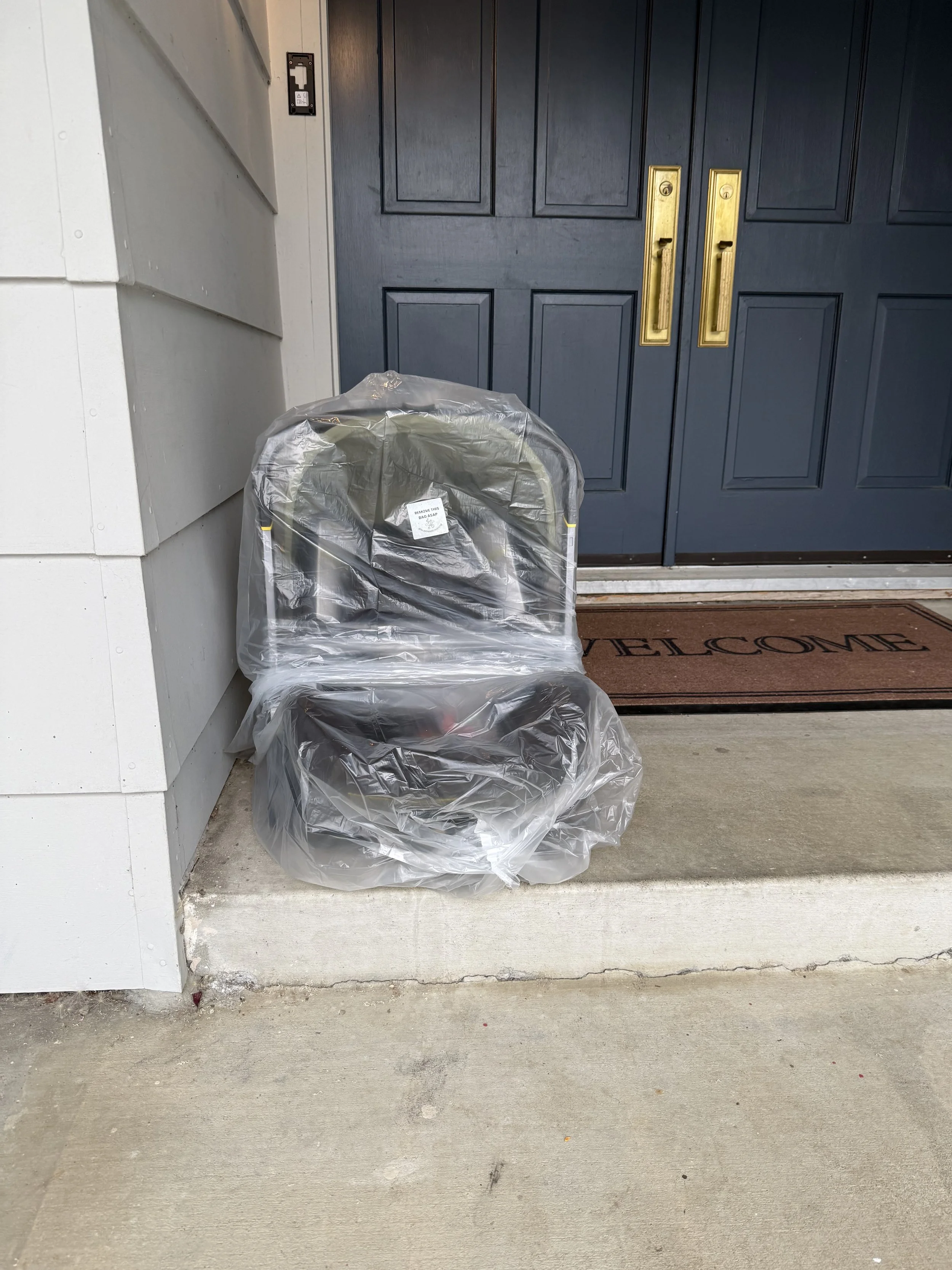  A plastic covered car seat sits on the ground outside a house next to a blue double door with gold handles. A brown doormat with black lettering that reads 'WELCOME' is in front of the door.