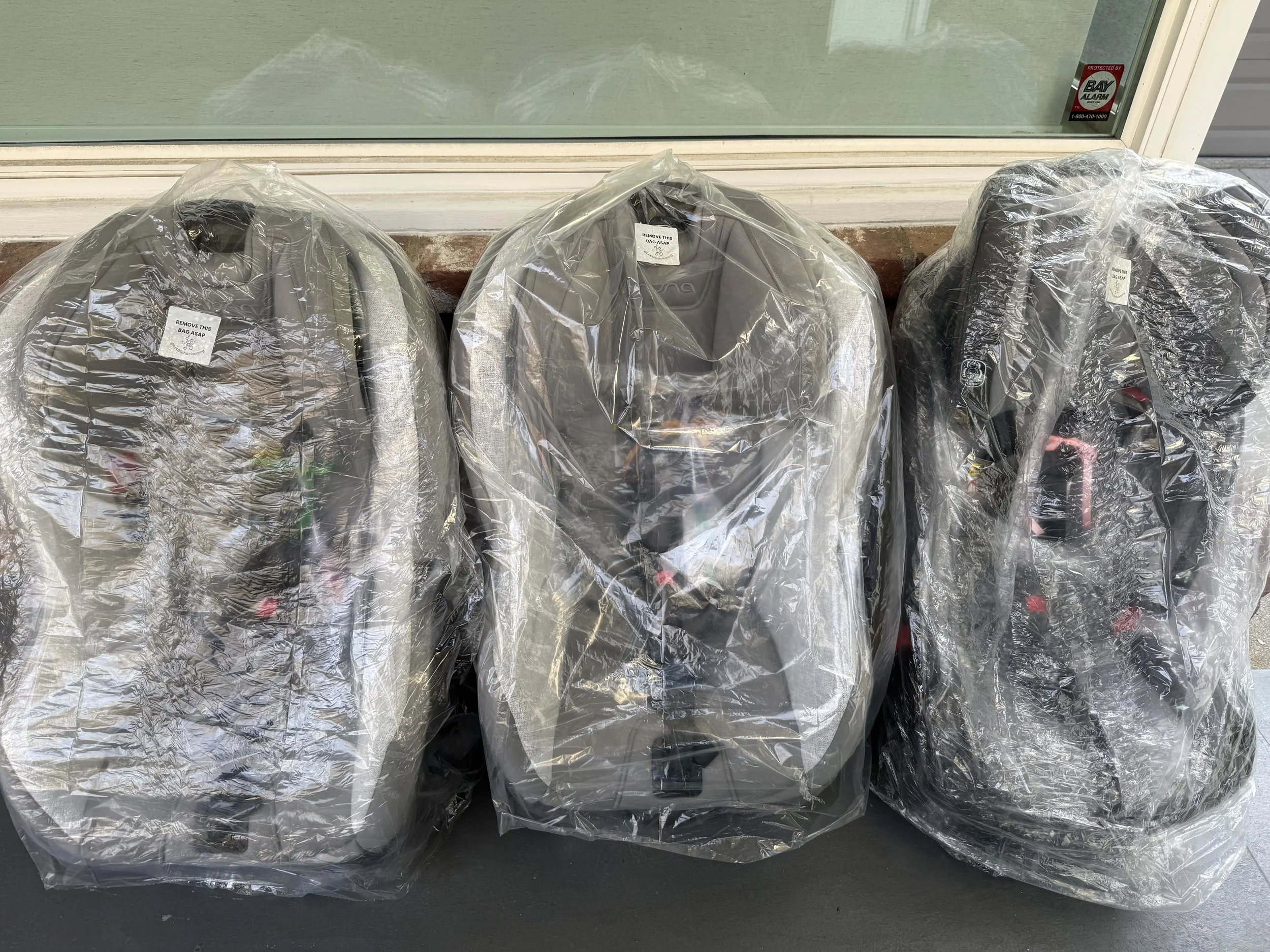 Three car seats wrapped in clear plastic bags, placed on the ground in front of a glass door with a security sticker.