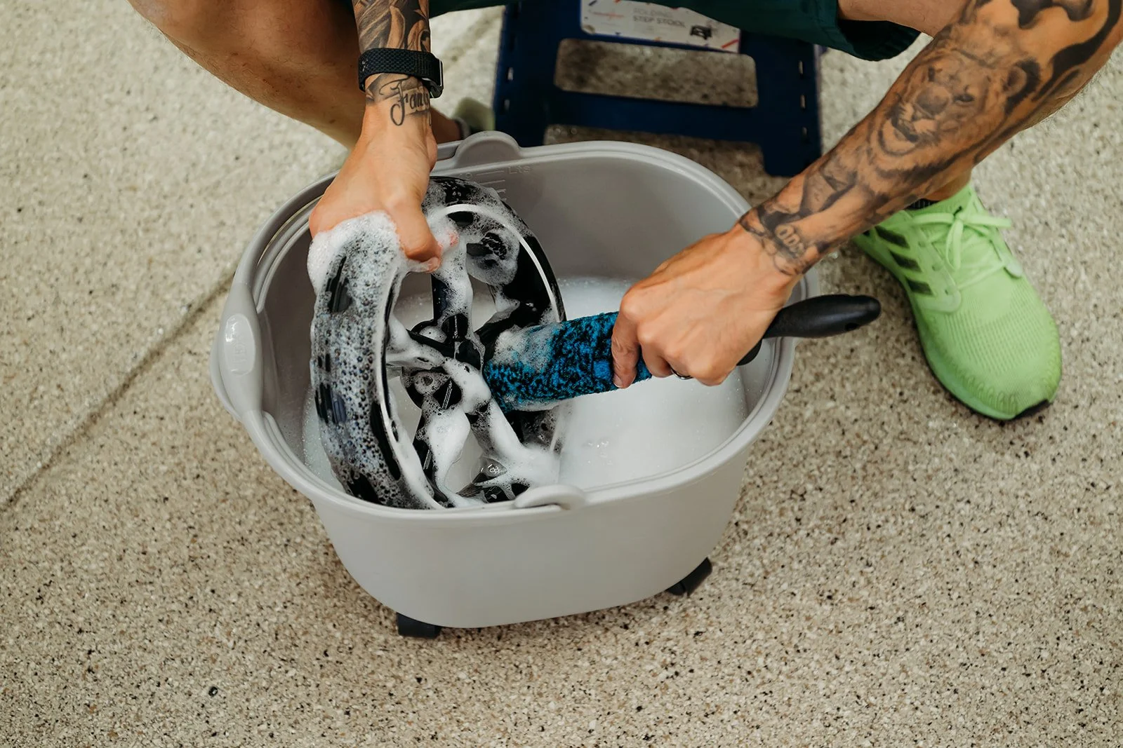 Man washing a pair of stroller wheel in a grey bucket filled with soap suds, on a beige garage floor.