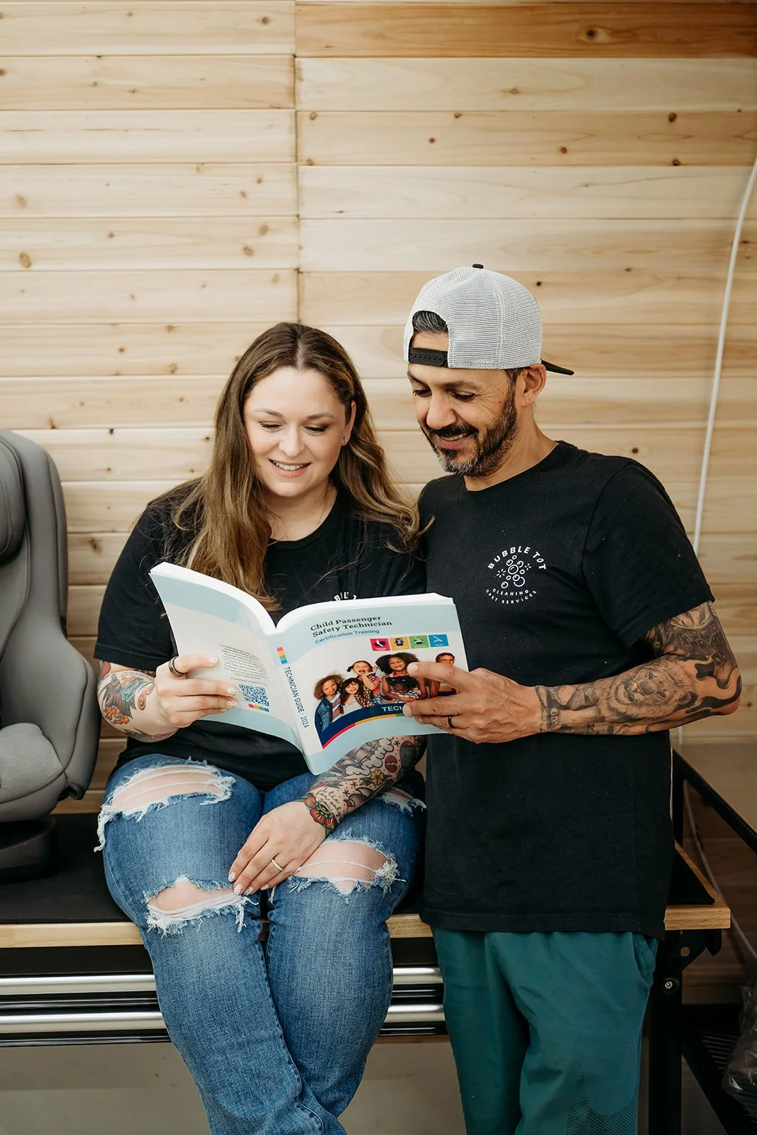 A woman and a man look at a booklet titled 'Child Passenger Safety Technician Certification Training' together in a room with wood-paneled walls. Both are wearing black t-shirts, and the woman has tattoos on her arms and ripped jeans.