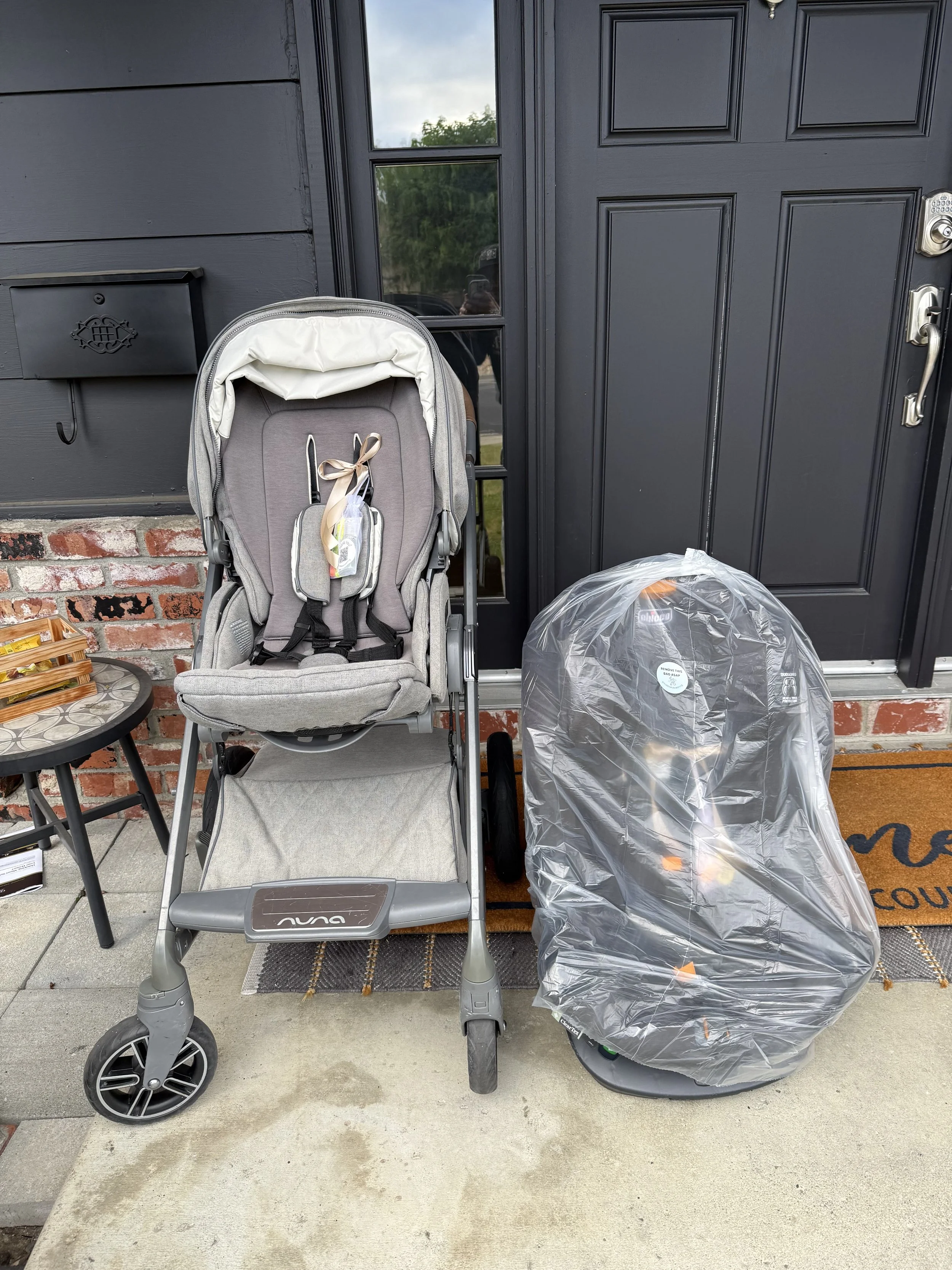 Front door with a gray baby stroller and a covered car seat on the porch, including a small hanging basket, a mailbox, and a doormat with black text, brick steps, and a concrete porch.