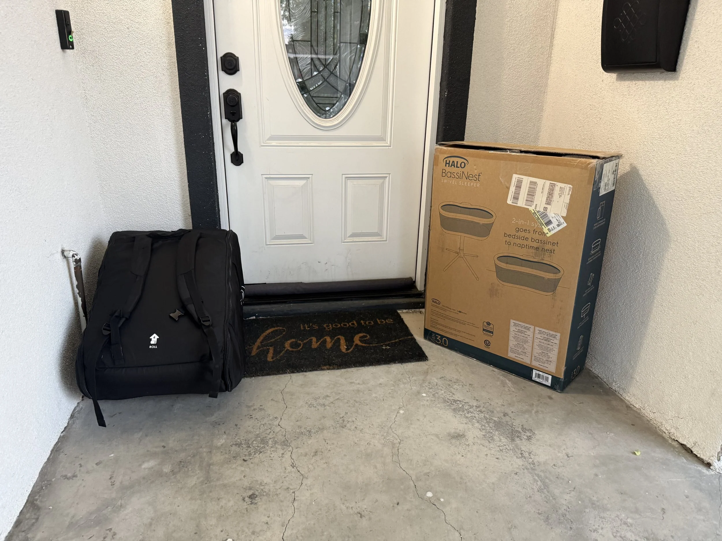 Front door with a black handle and keypad lock, a large cardboard box with product details of a HALO BassiNest, a black Doona brand bag on the floor, a welcome mat, and a small black mailbox on the wall.