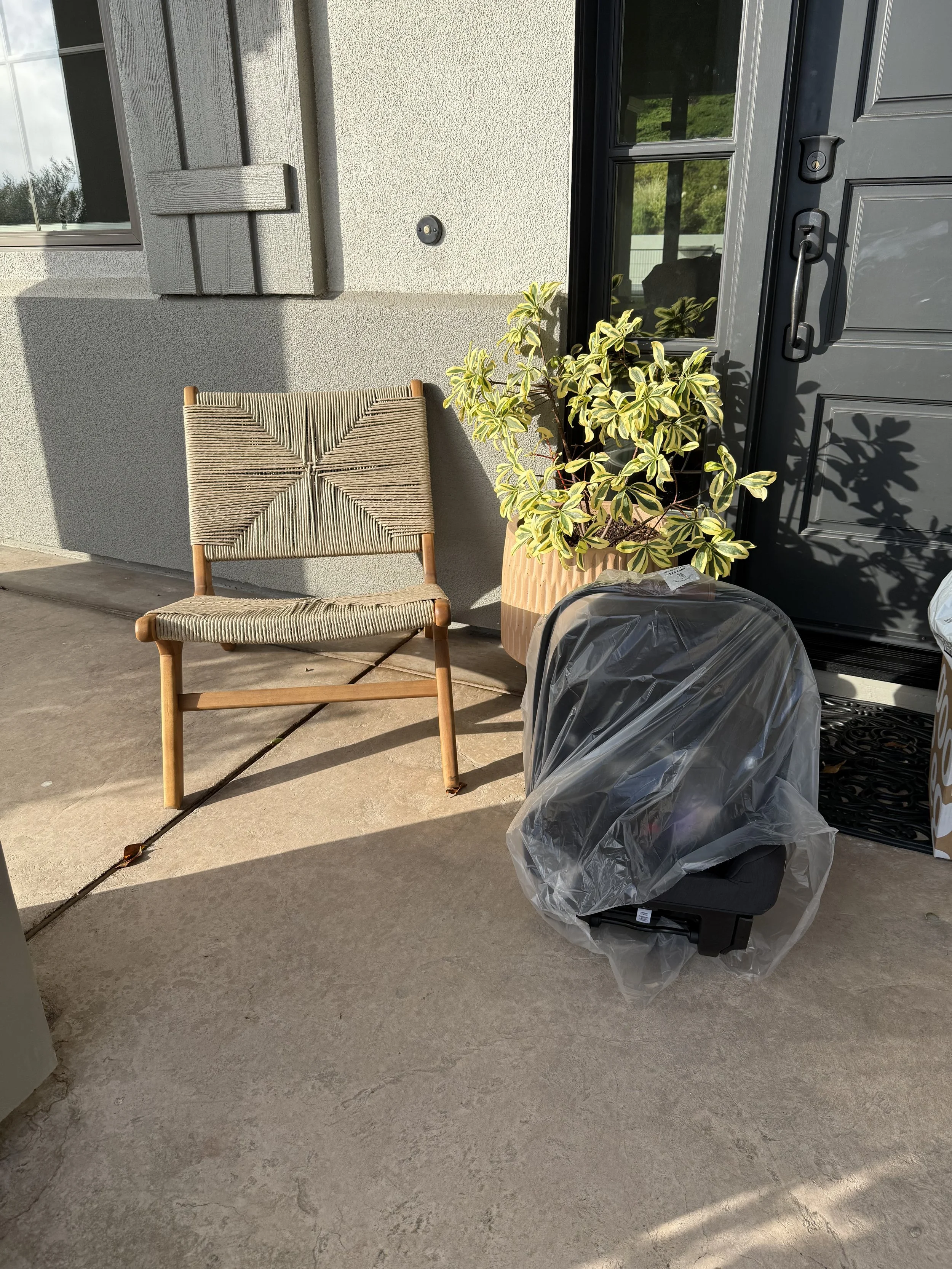 A porch area with a wicker chair, a potted plant with variegated leaves, a covered car seat and a black door with decorative molding. The porch has a concrete floor and part of a window is visible on the left side.