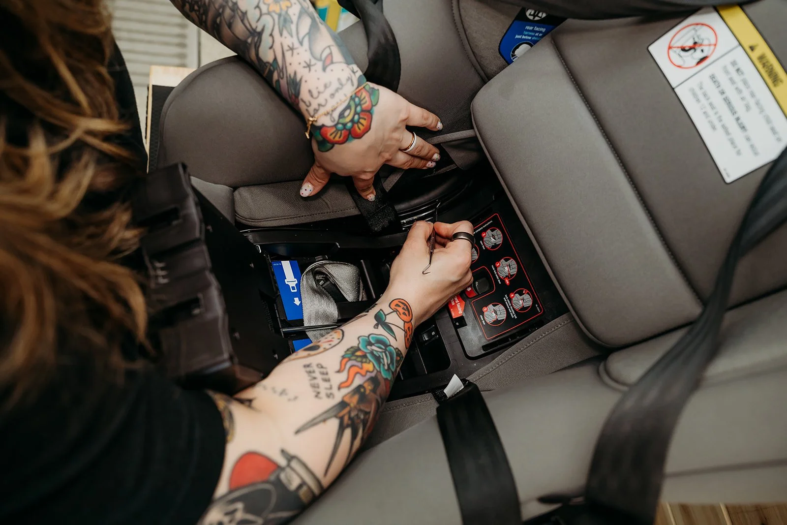 A person with tattoos on their arms, including a colorful flower and the phrase 'Never Sleep', is cleaning a car seat. She is using a picker tool to pick at the crevices of the car seat.