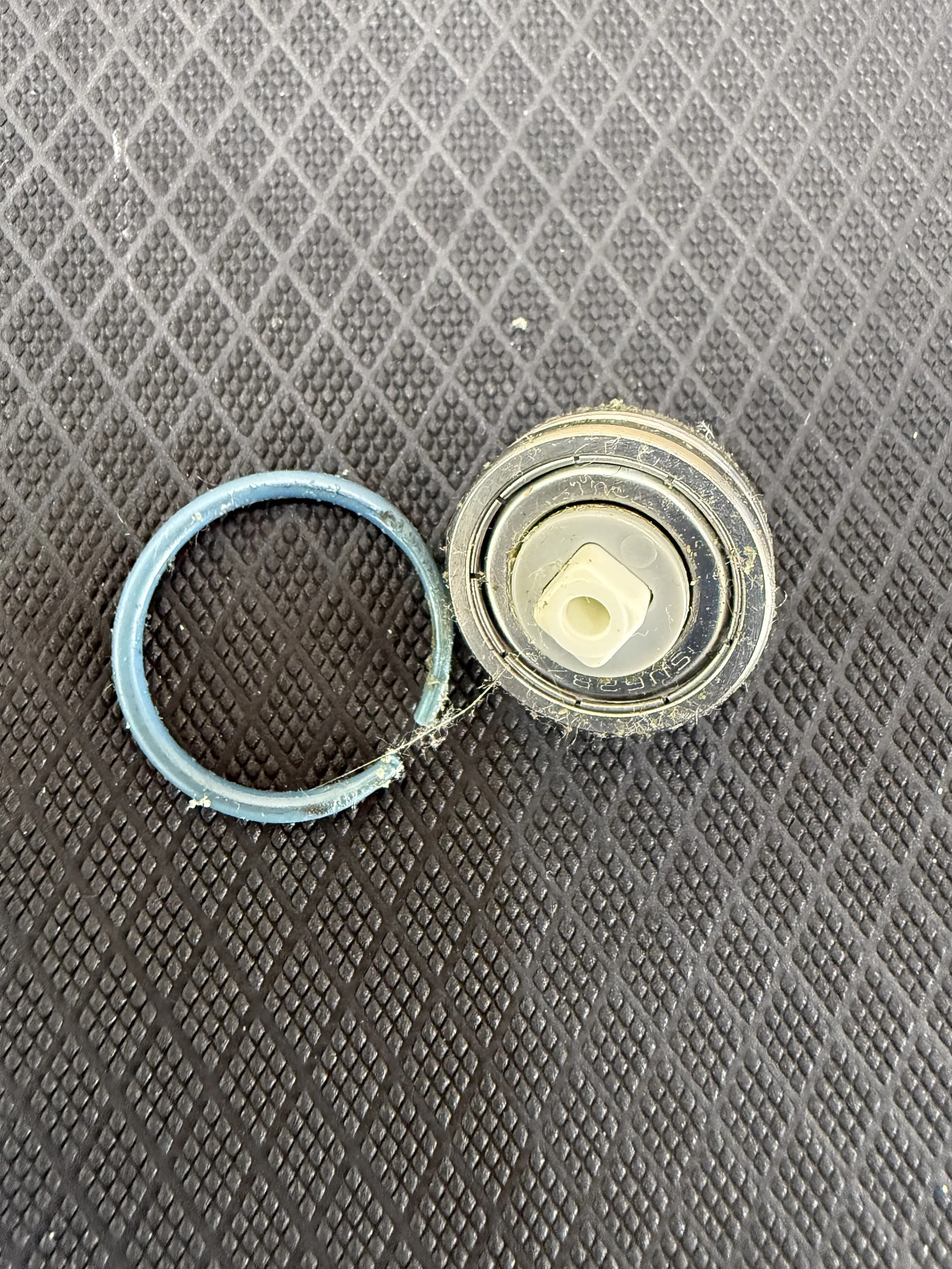 A broken SNOO o-ring bearing with a circular blue rubber gasket and a metal cover with a plastic or ceramic core on a textured black rubber mat.