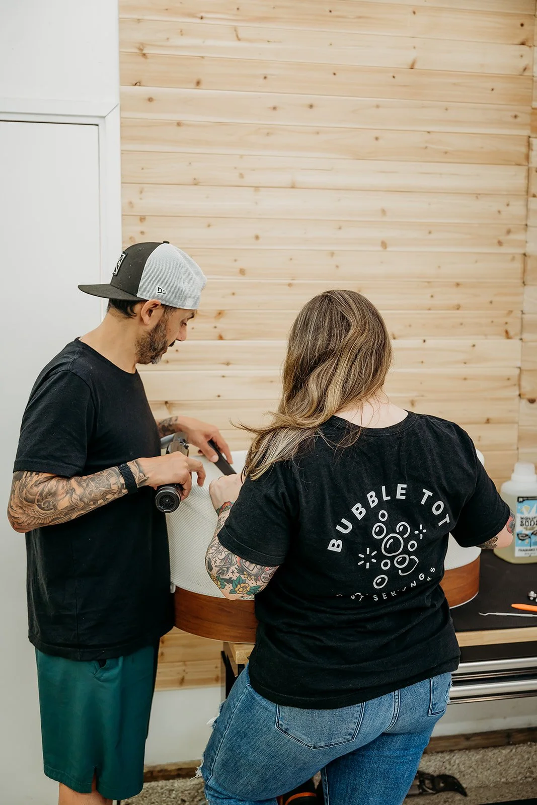 A man and a woman with tattoos working together on a project in a room with wood-paneled walls. The man is holding a tool, and the woman is wearing a black t-shirt with a bubble design on the back. There are supplies on a table nearby.