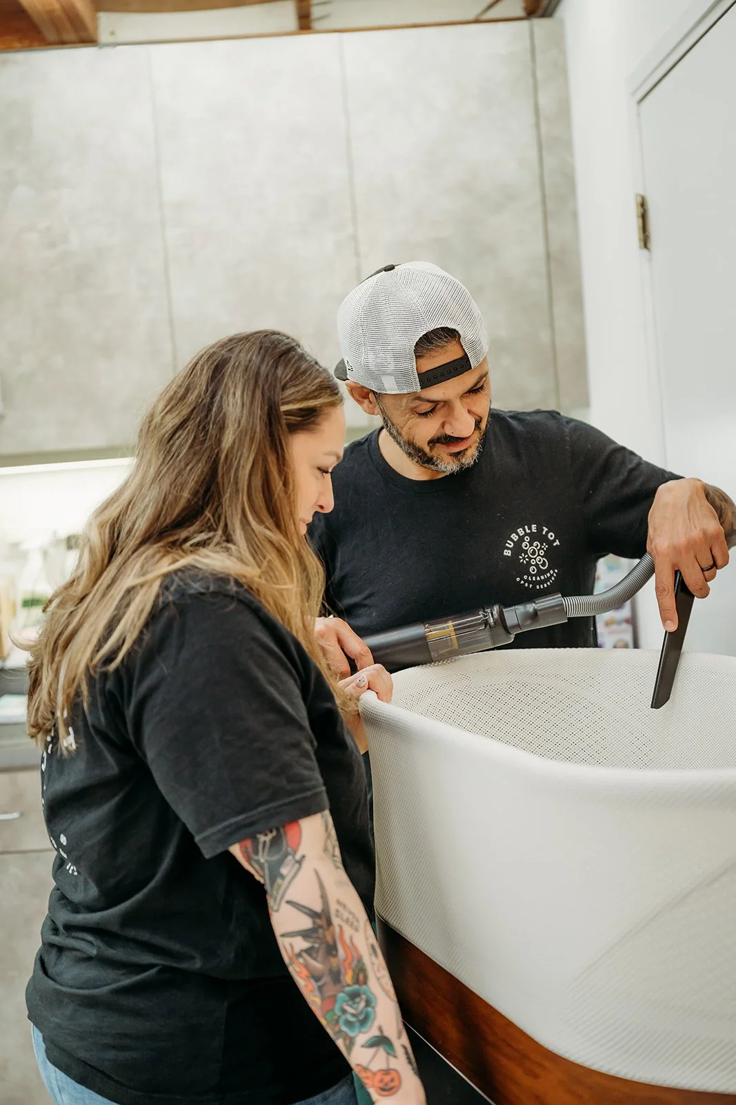 A man and a woman are cleaning a SNOO together with a handheld vacuum cleaner. The man is wearing a black t-shirt and a backwards white baseball cap, while the woman has long, tattooed arms and is wearing a black t-shirt.