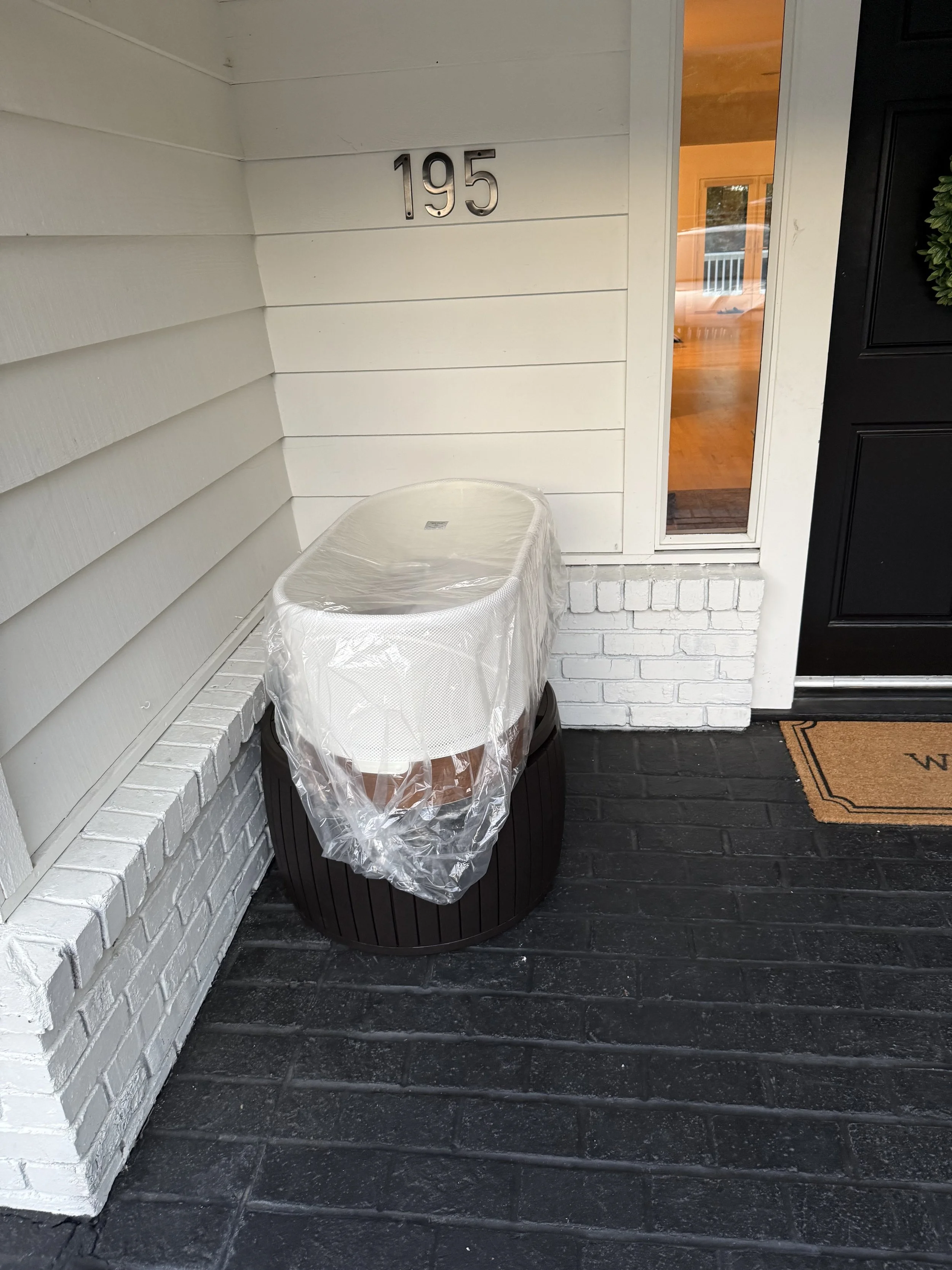 Front porch with house number 195, a black door with a small glass window, white siding, a white brick part, a black wooden planter, and a mat with a decorative border. A covered SNOO is wrapped in plastic and placed on a black wooden stand.