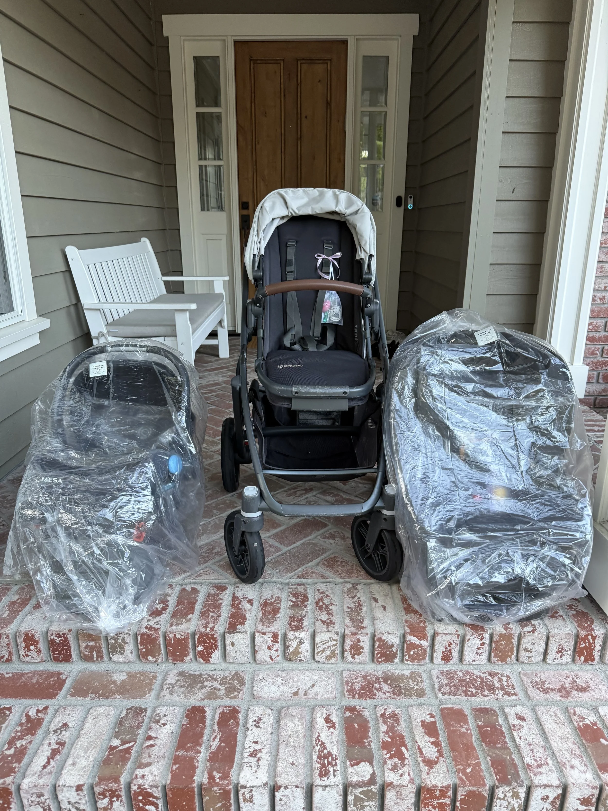 A front porch with a baby stroller in the center, two covered car seats on either side, a white bench to the left, and a wooden door with glass panels behind the stroller. The porch has a brick floor and is surrounded by siding walls.