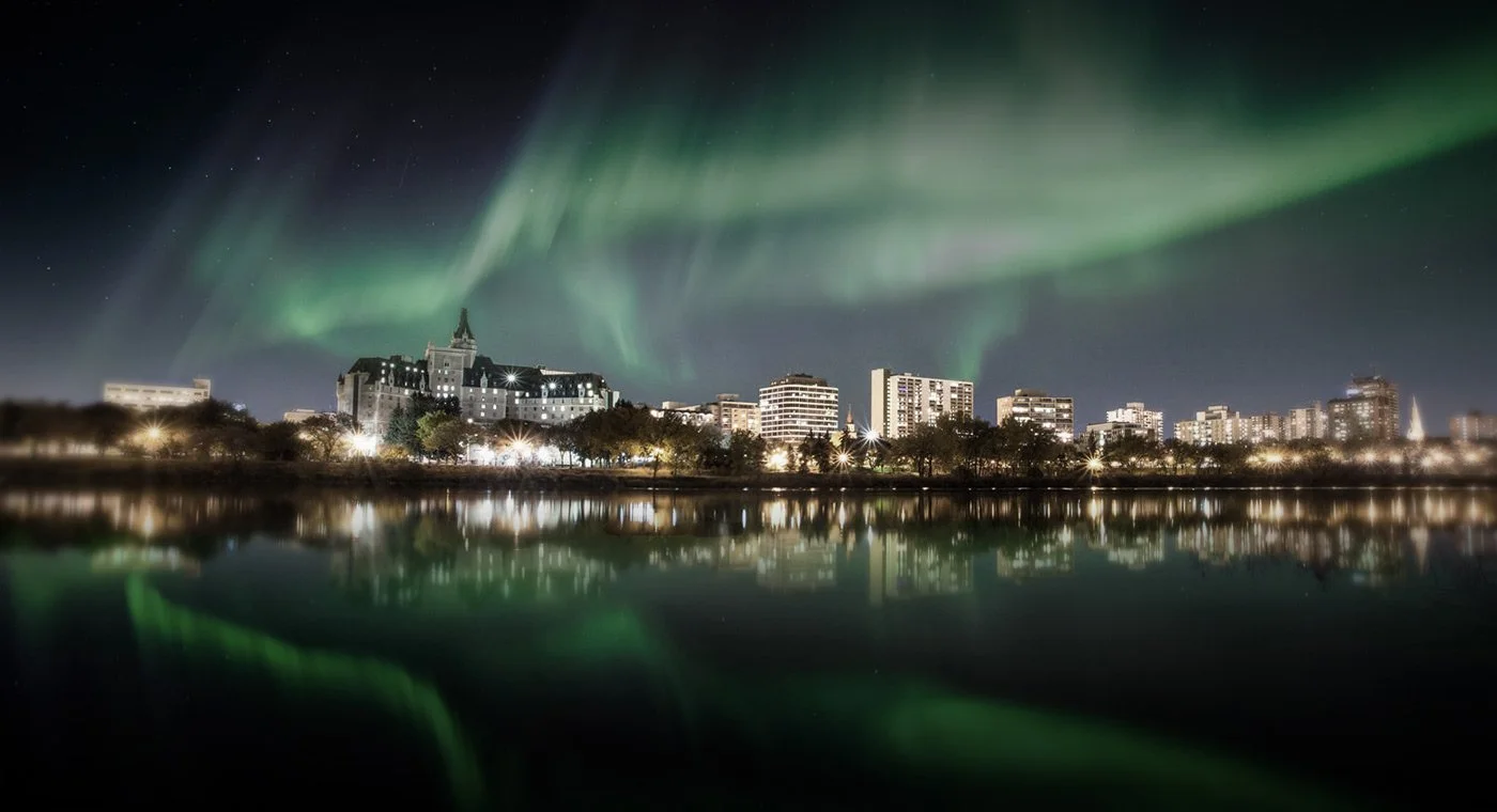Nighttime cityscape with illuminated buildings and a lake reflecting the city lights, with green northern lights or aurora borealis in the sky above.