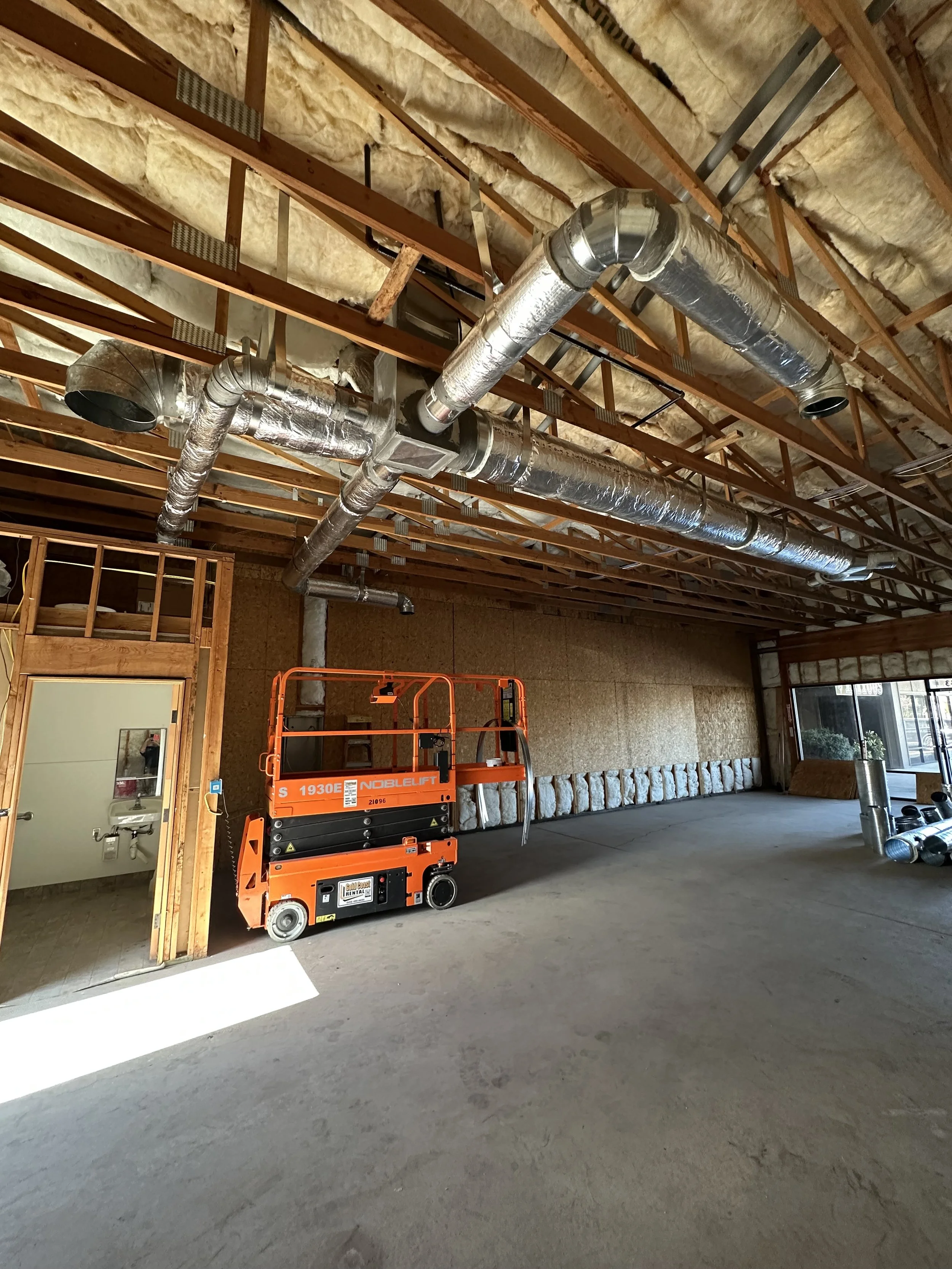 Construction site interior with exposed ceiling joists, ductwork, insulation, and an orange lift.