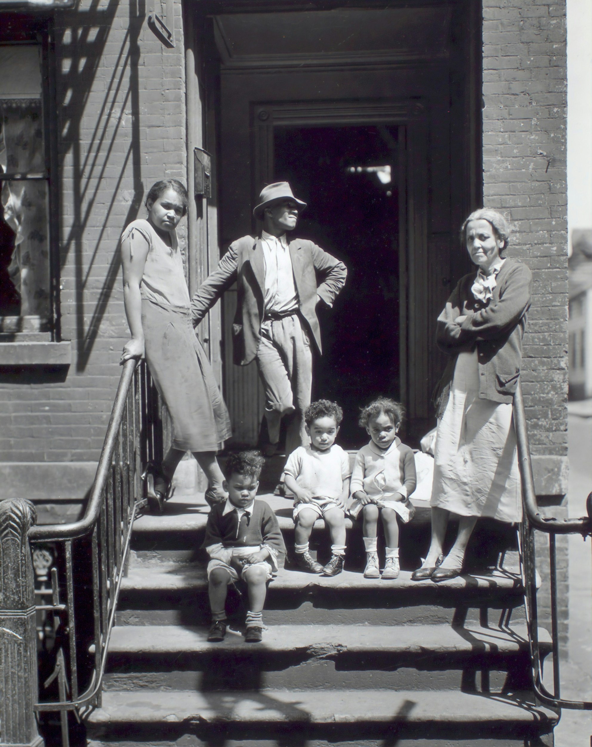 Black and white photograph of three children sitting on steps and two women standing nearby in front of a brick building, with one man standing in the doorway.