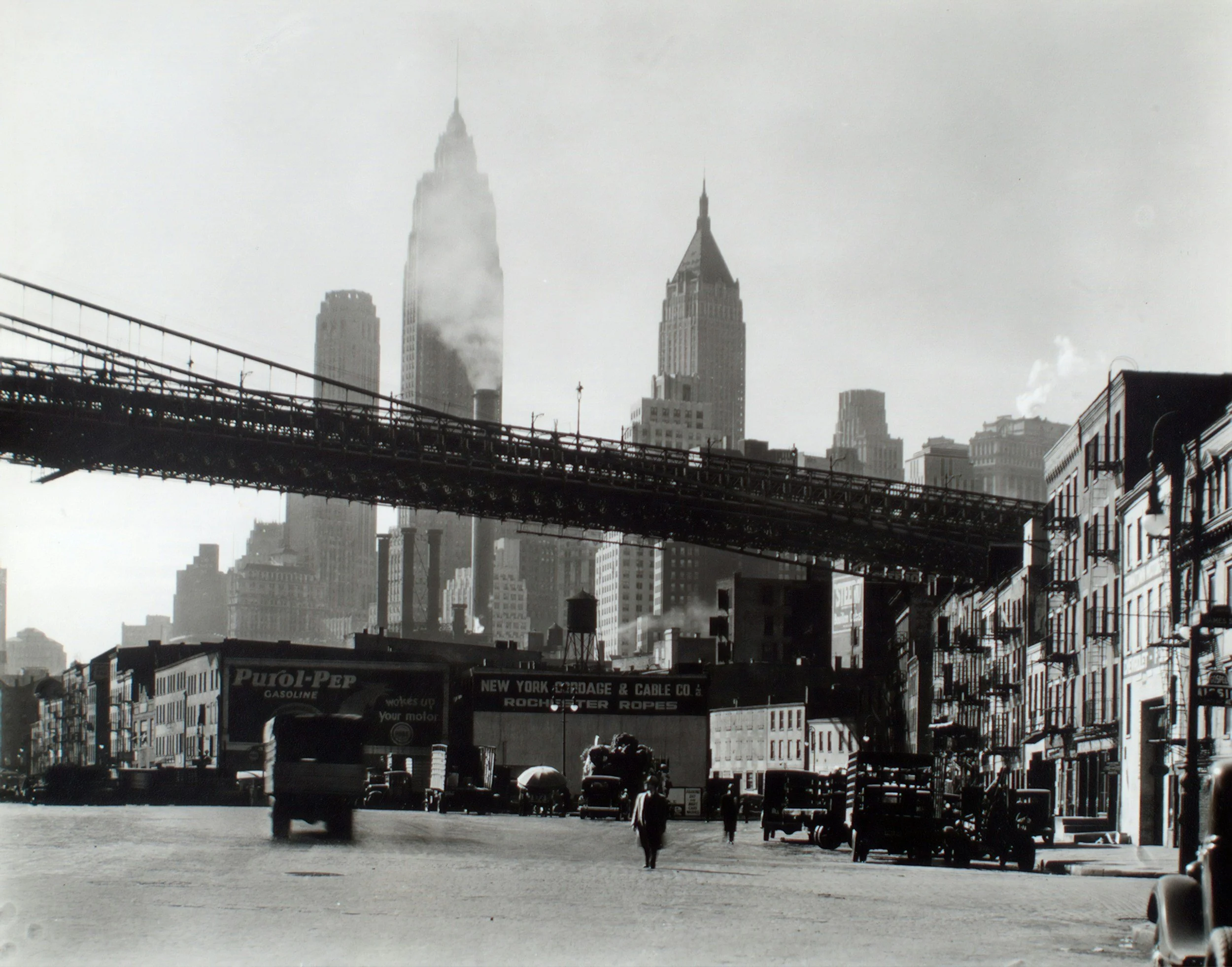 A black-and-white photo of a city street view with the Empire State Building in the background, an elevated train track running across the scene, and people walking along the sidewalk, with old-style cars parked on the street.