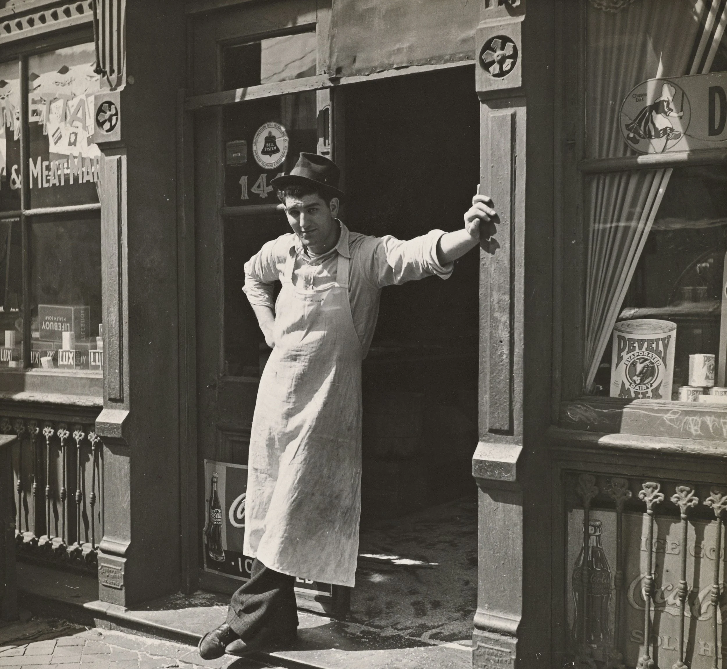 A young man wearing a hat and apron standing in the doorway of a shop, leaning against the doorframe with one hand on his hip and the other on the doorframe, smiling.
