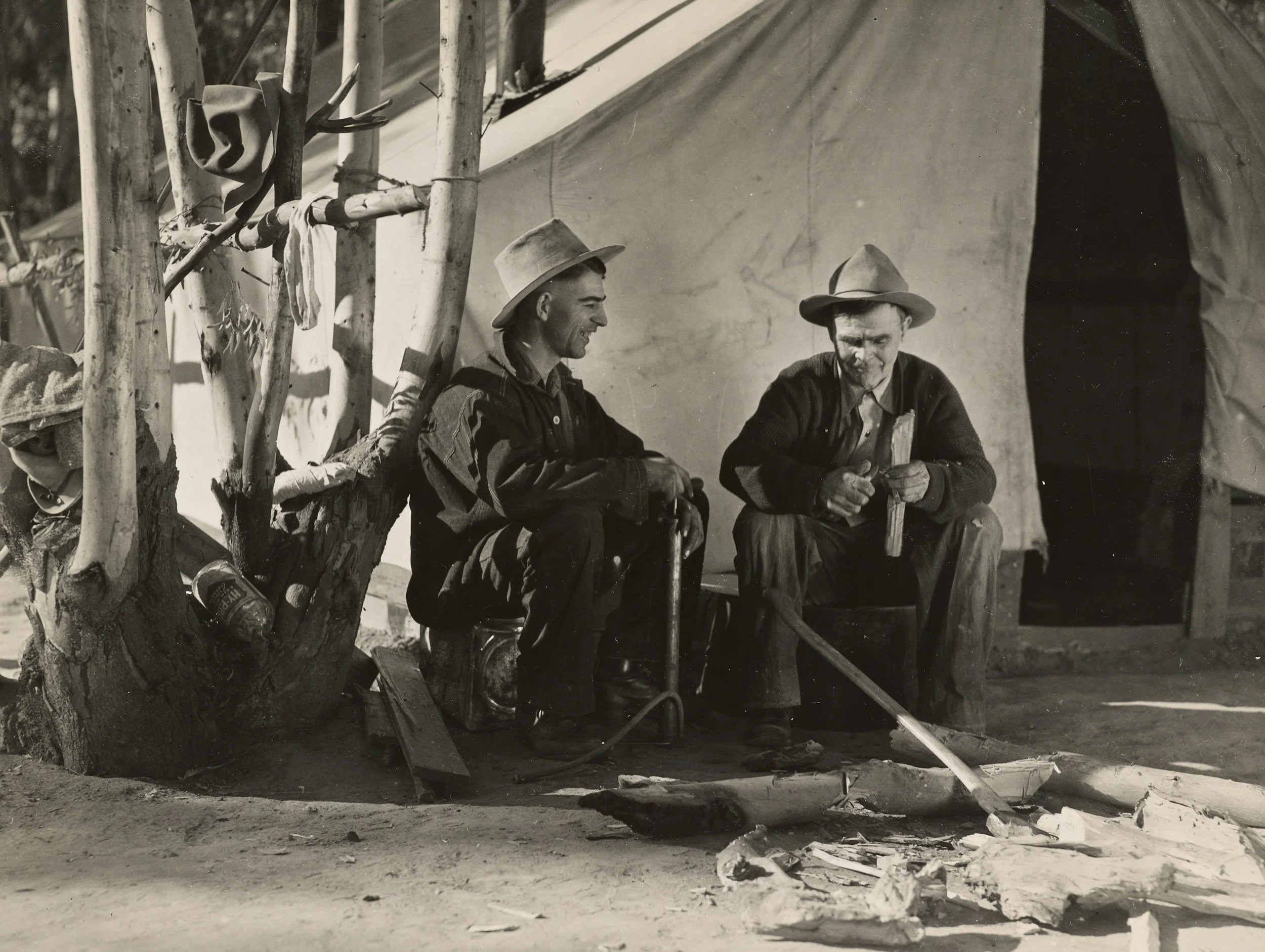 Two men wearing hats sitting outside a tent, with one carving wood and the other smiling.