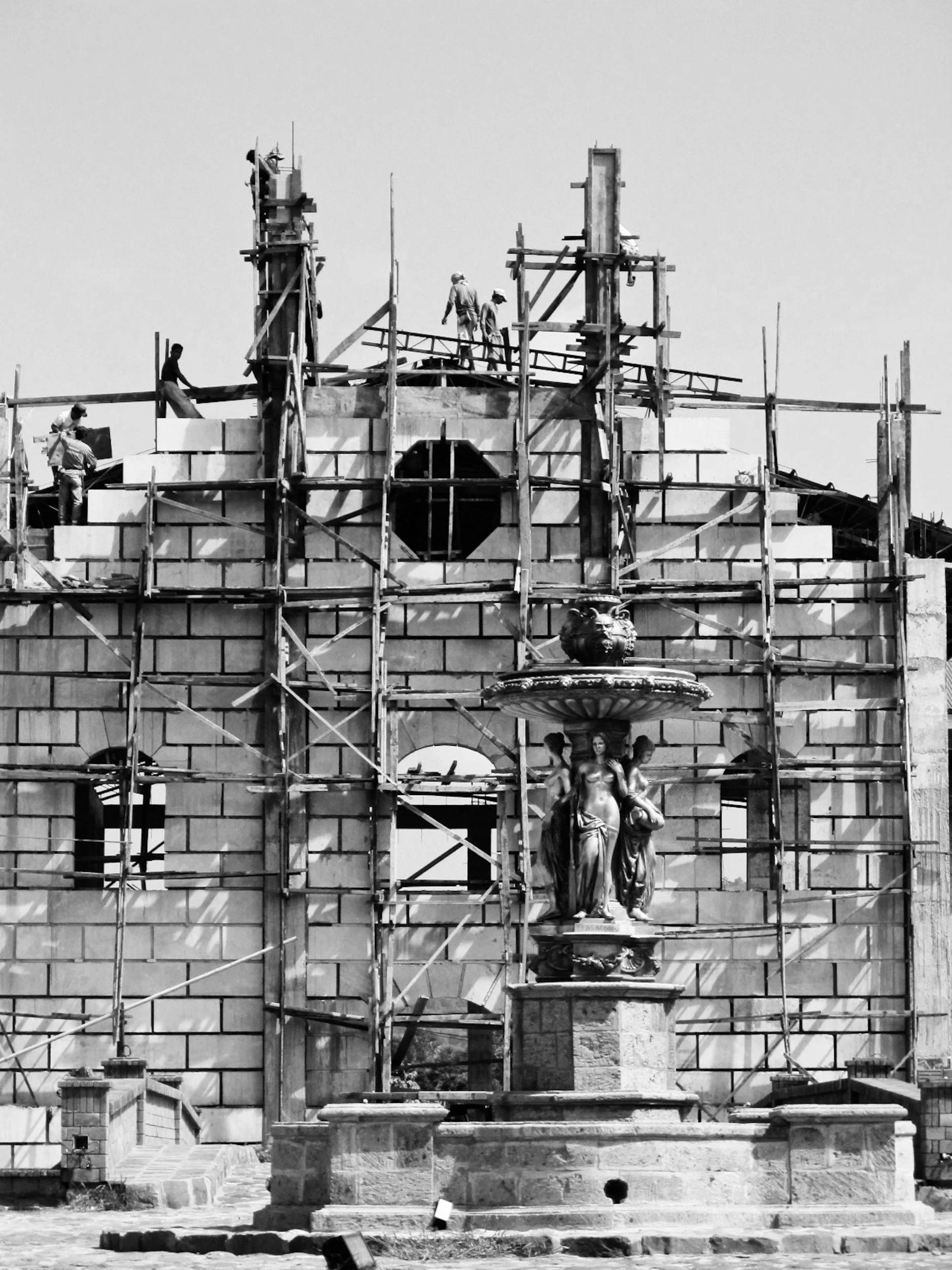 Construction workers working on a building with scaffolding, and a fountain with statues in the foreground.