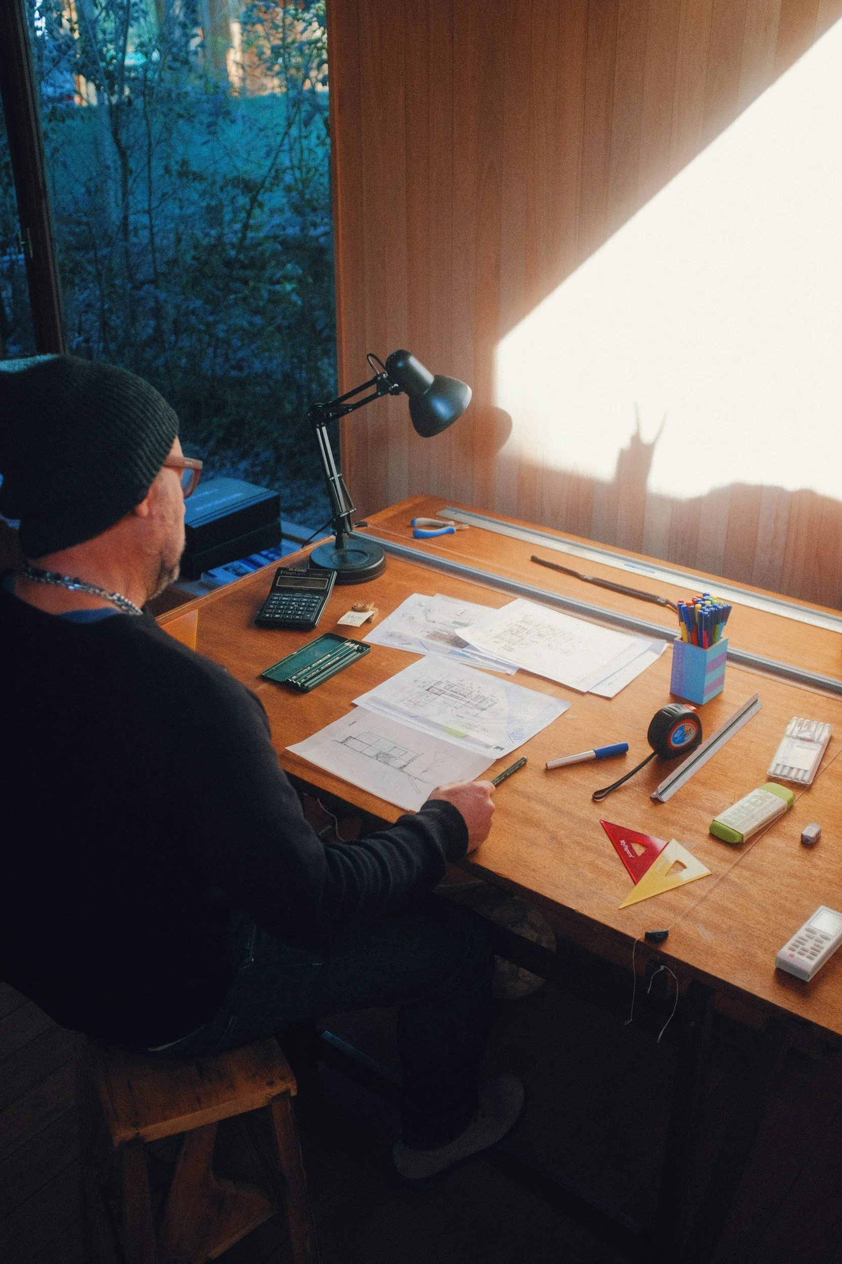 A person sitting at a wooden desk with blueprints and drawing tools, illuminated by sunlight coming through a window with a view of trees outside.