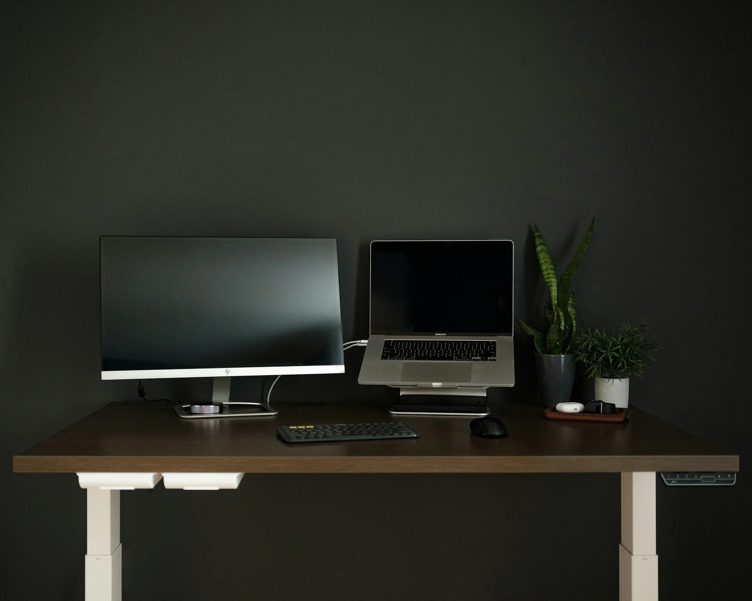 A modern workspace with a wooden desk against a dark wall, featuring a desktop computer with a large monitor, a laptop on a stand, a wireless keyboard and mouse, two potted plants, a wireless earbud case, a smartwatch, and a small tray with watches.