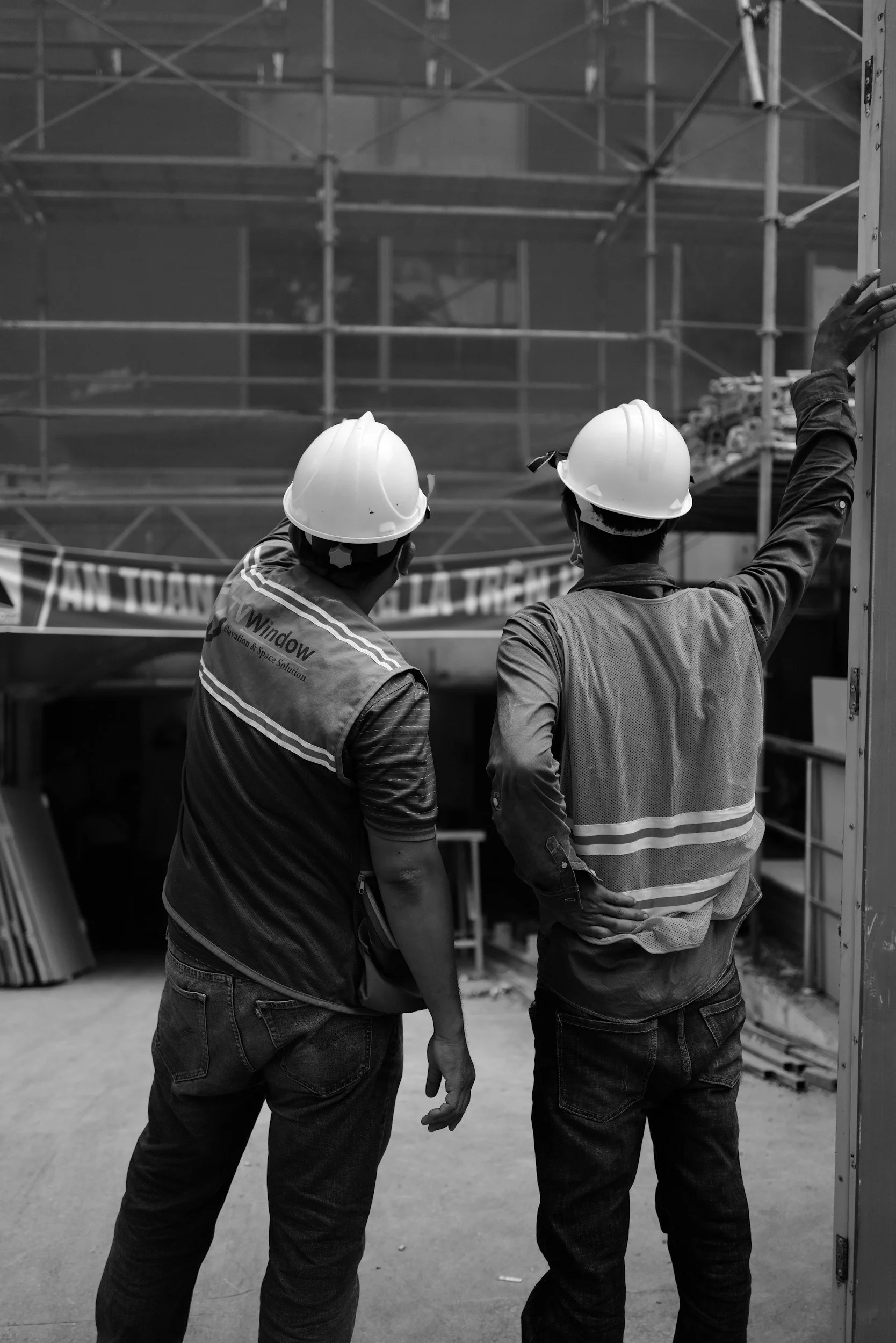 Two construction workers wearing helmets and safety vests discussing a construction site with scaffolding.