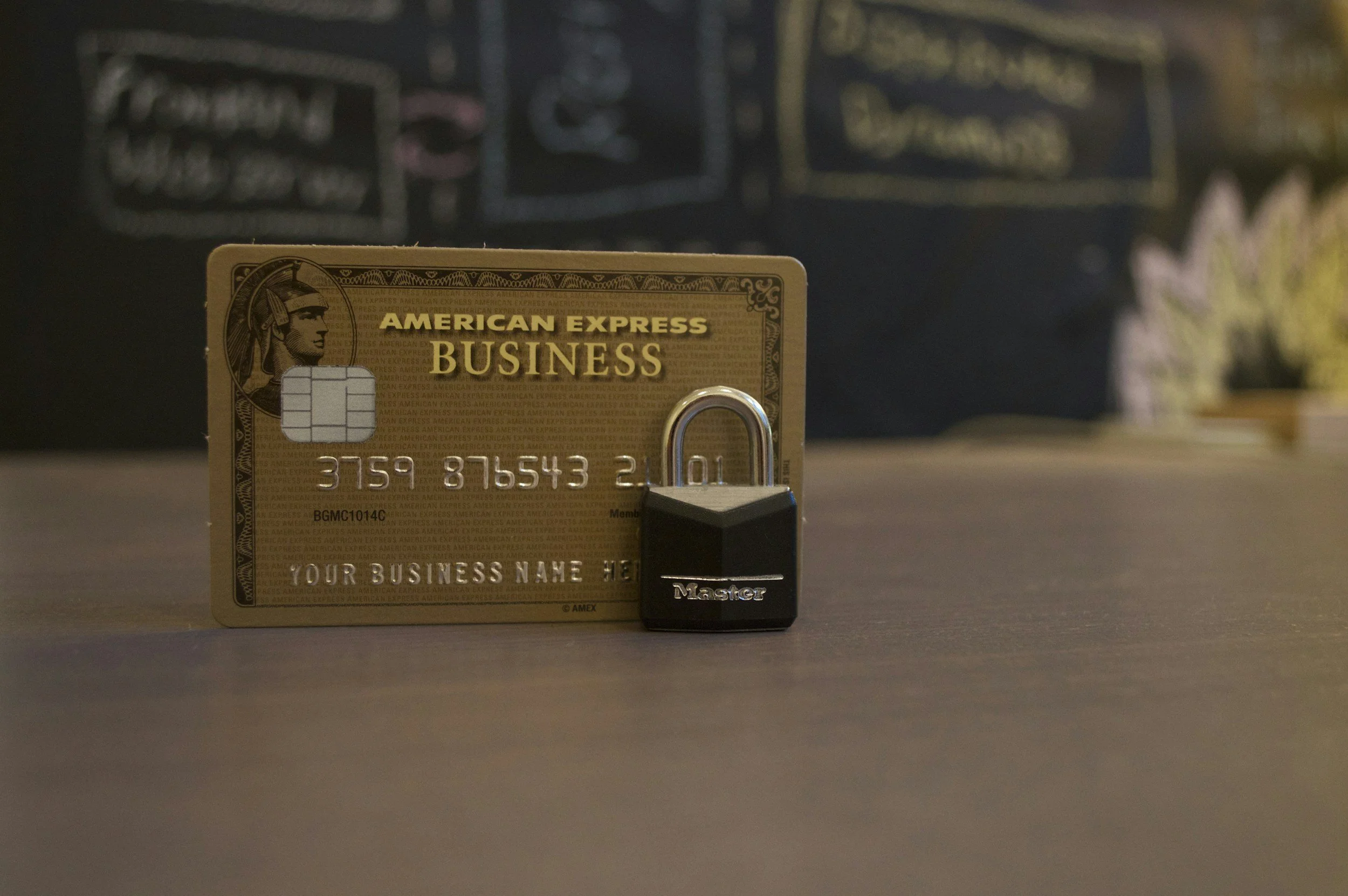 Gold American Express Business credit card with a black Master lock in front of it, placed on a wooden surface.