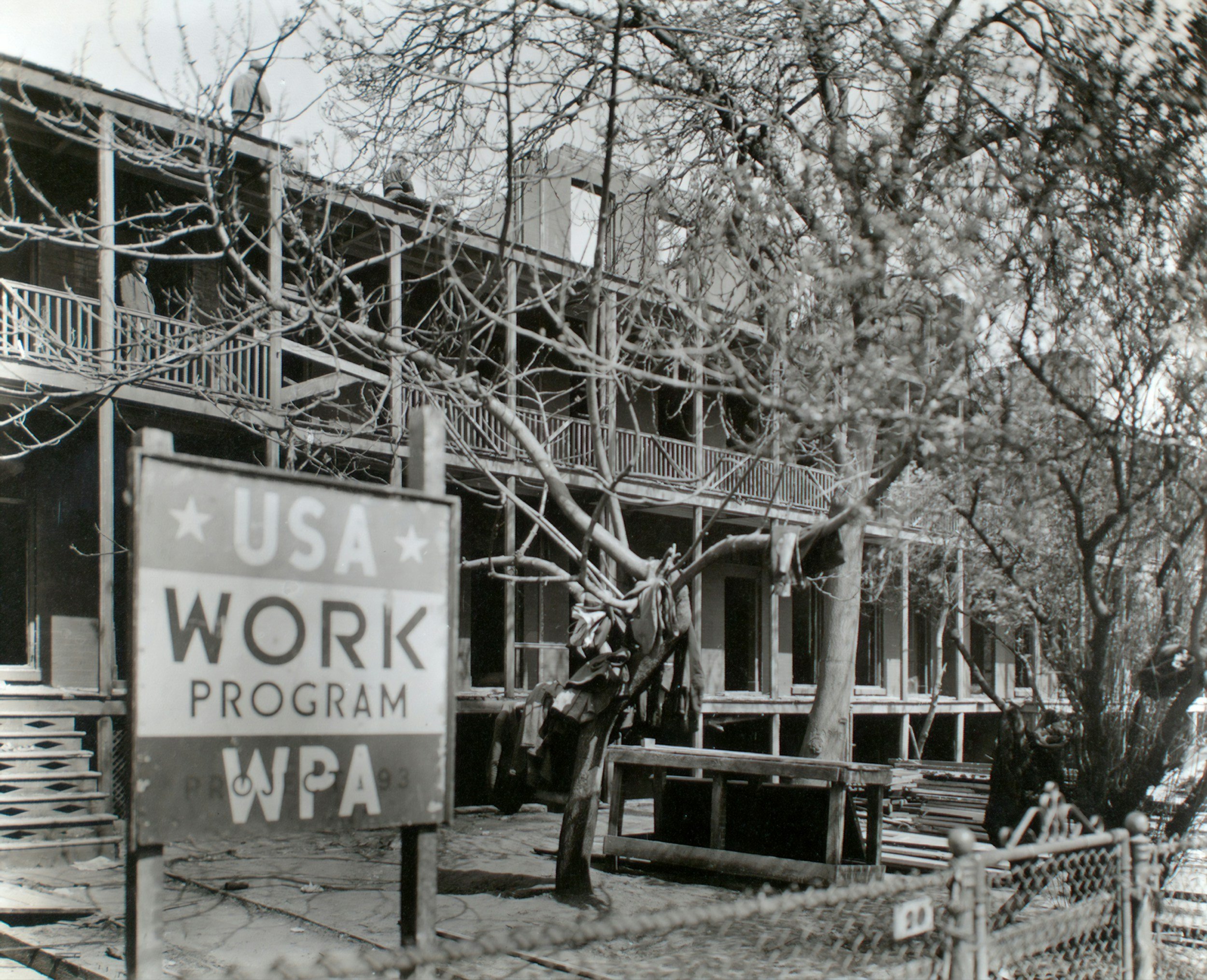 Black and white photo of a building with balconies, surrounded by leafless trees, with a sign in the foreground reading 'USA WORK PROGRAM WPA'.