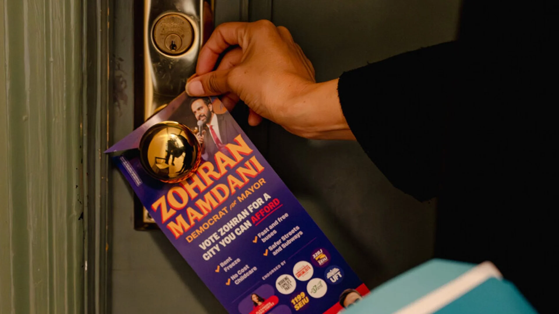 Person leaving a campaign flyer on a door for Zohran Mamdani, a Democratic candidate for mayor, promoting his policies on rent freeze, no cost childcare, and affordability in the city.