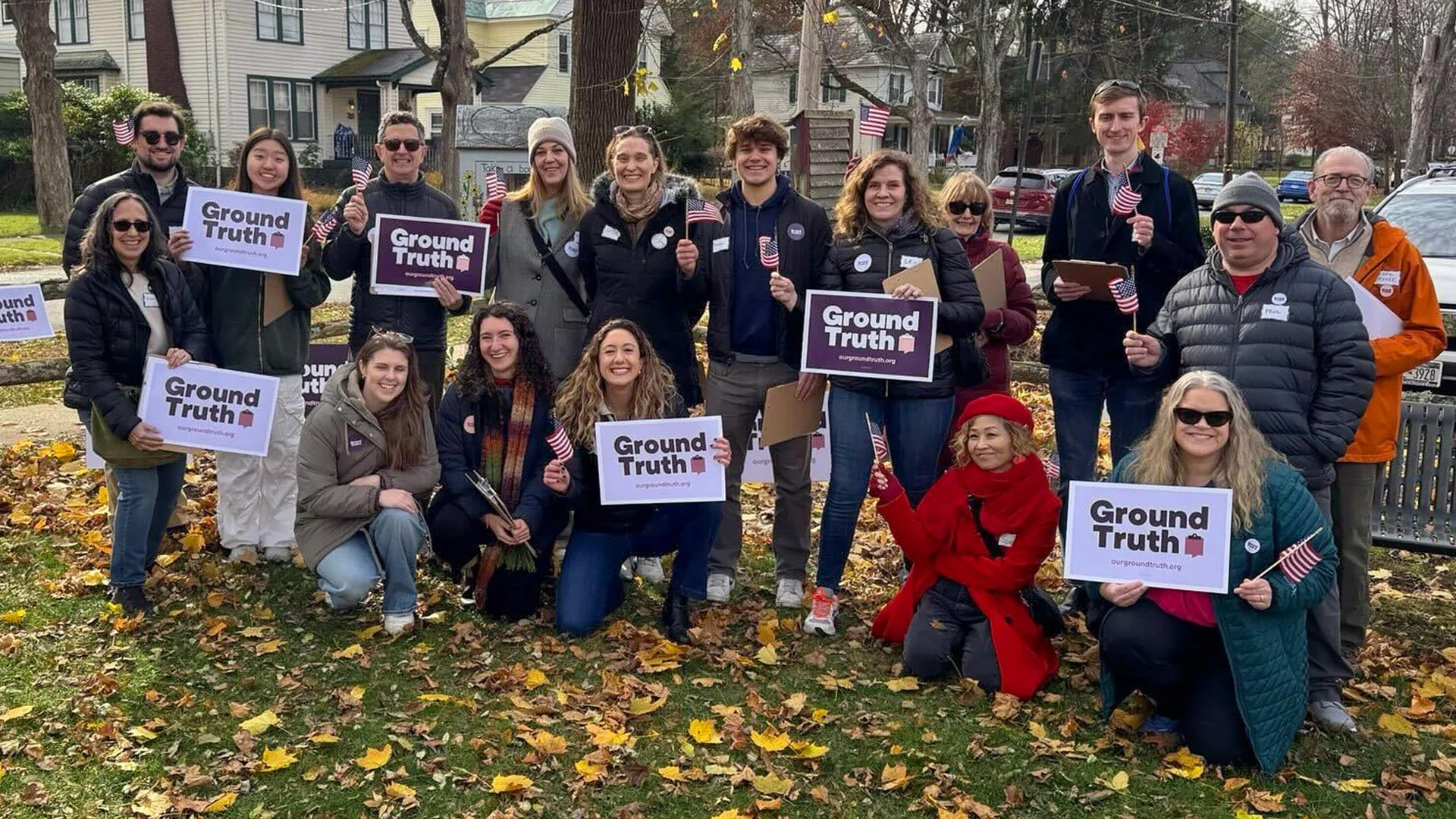Group of people gathered outdoors during fall, holding 'Ground Truth' signs and small American flags, smiling for a photo.