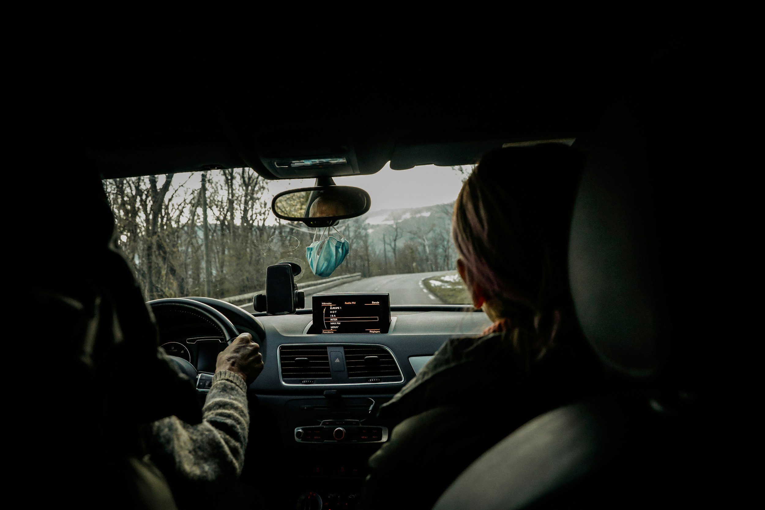Inside a vehicle with two passengers, one driving and one in the front passenger seat, on a road with leafless trees on the sides during daytime.