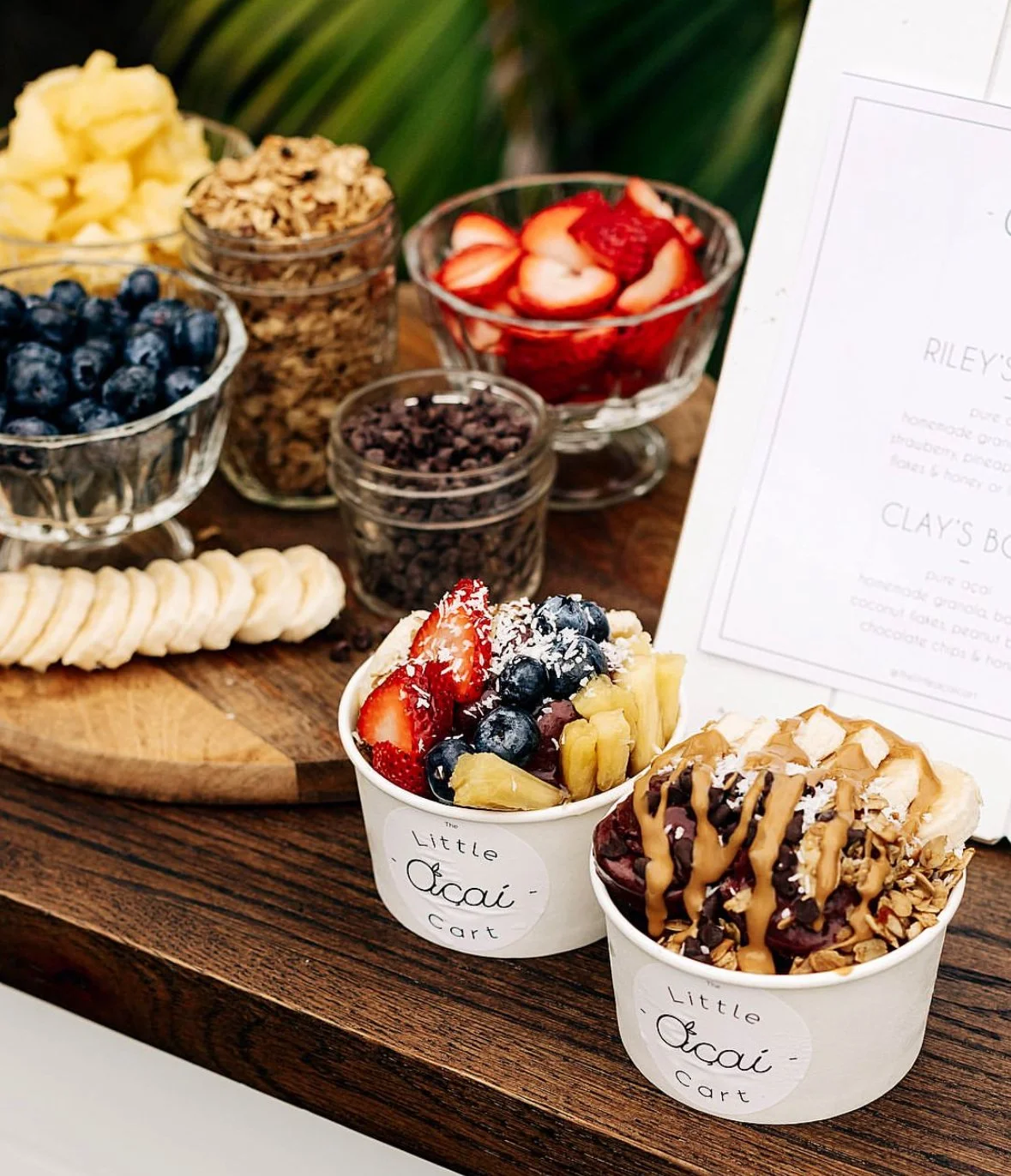 Assorted bowls and containers of fresh fruit, gluten-free granola, and toppings on a wooden cart, including strawberries, blueberries, pineapple,  and chocolate peanut butter cups, with two different acai bowls decorated with fruit and toppings.