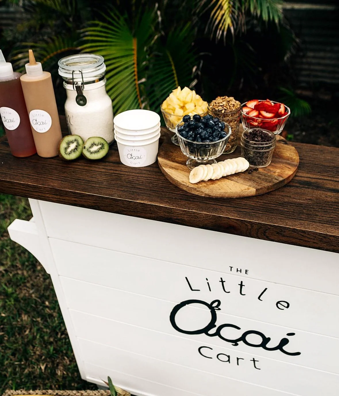 A wooden stand with a white sign that reads "The Little Ojai Cart," displaying an assortment of toppings and ingredients for acai bowls, including bottles, jars, sliced kiwi, blueberries, pineapple, strawberries, chopped chocolate, sliced banana, and granola, set outdoors with greenery in the background.