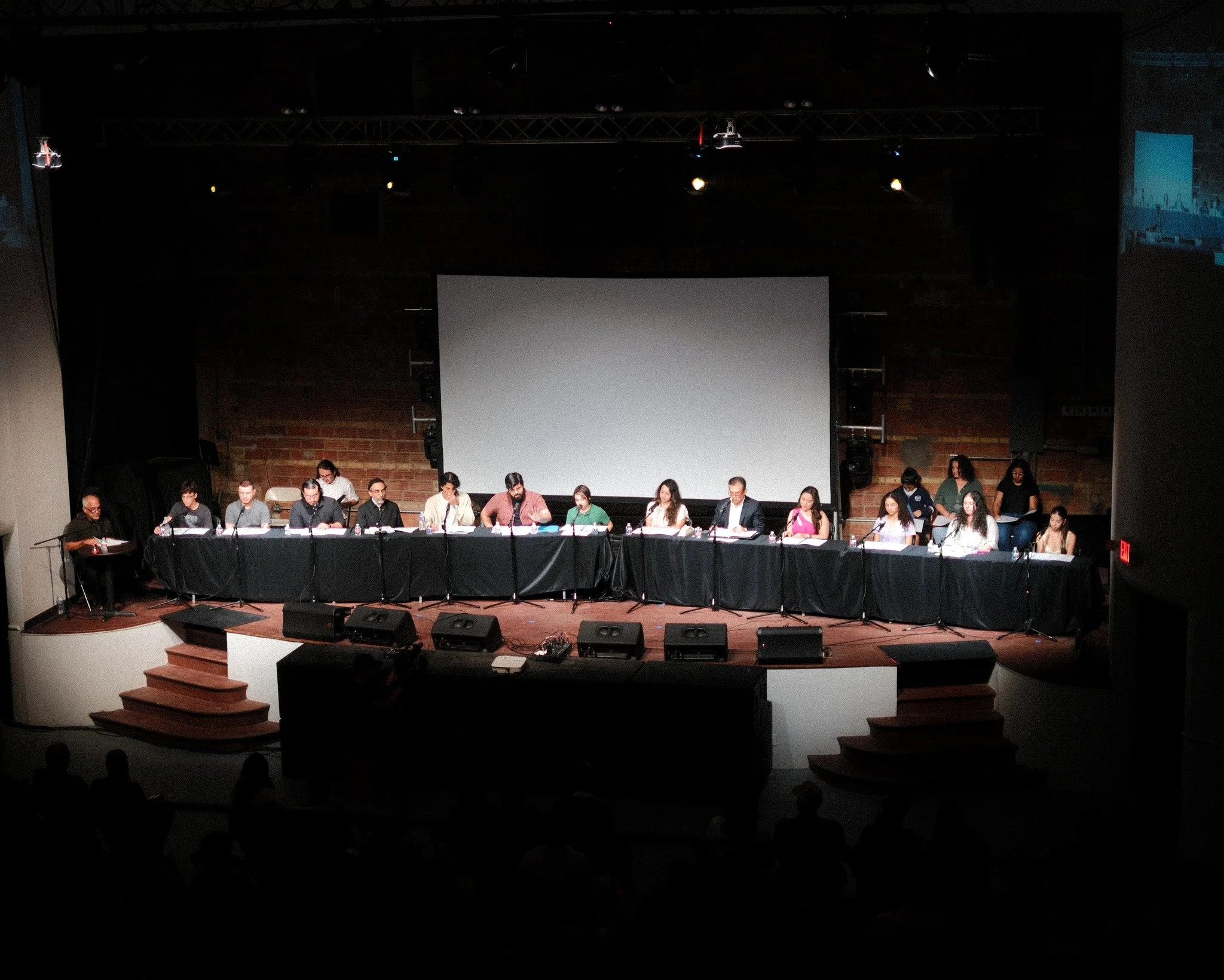 Panel of people seated at a long table on a stage during a conference or meeting, with a large blank screen behind them and a dark background.