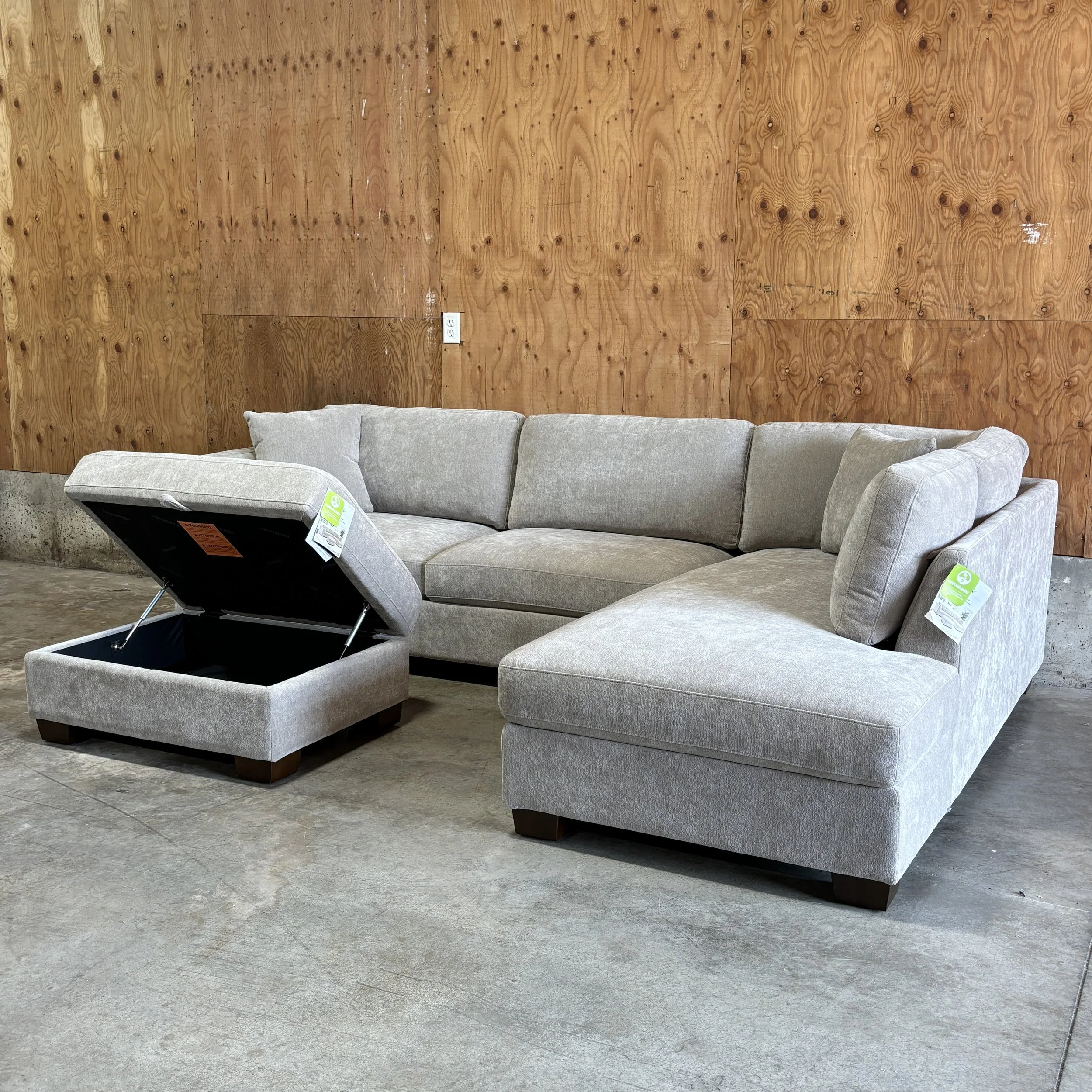Light gray sectional sofa with a matching ottoman in front of a wooden wall, with a concrete floor.