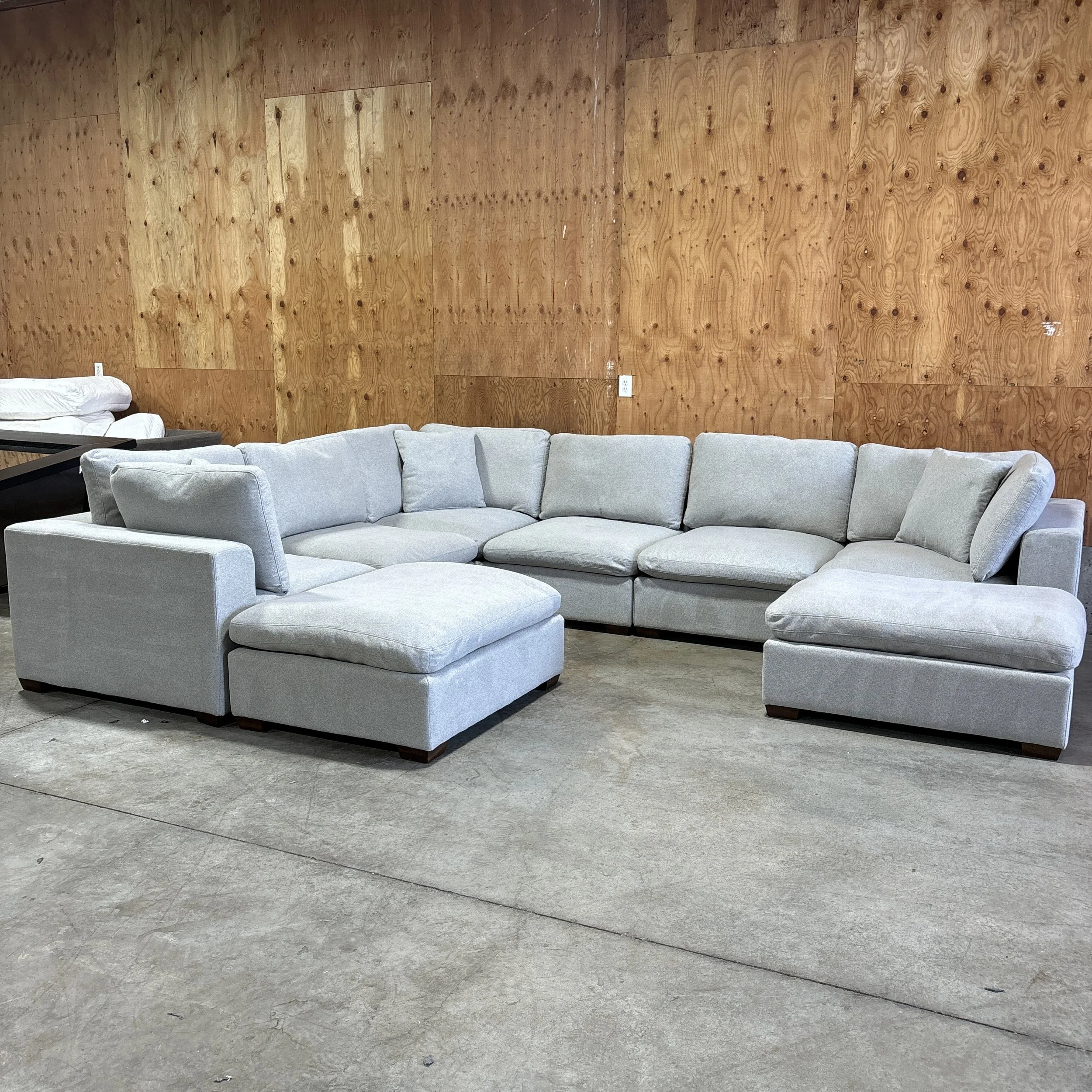 Light gray sectional sofa with cushions in front of a wood-paneled wall in a room with concrete floor.