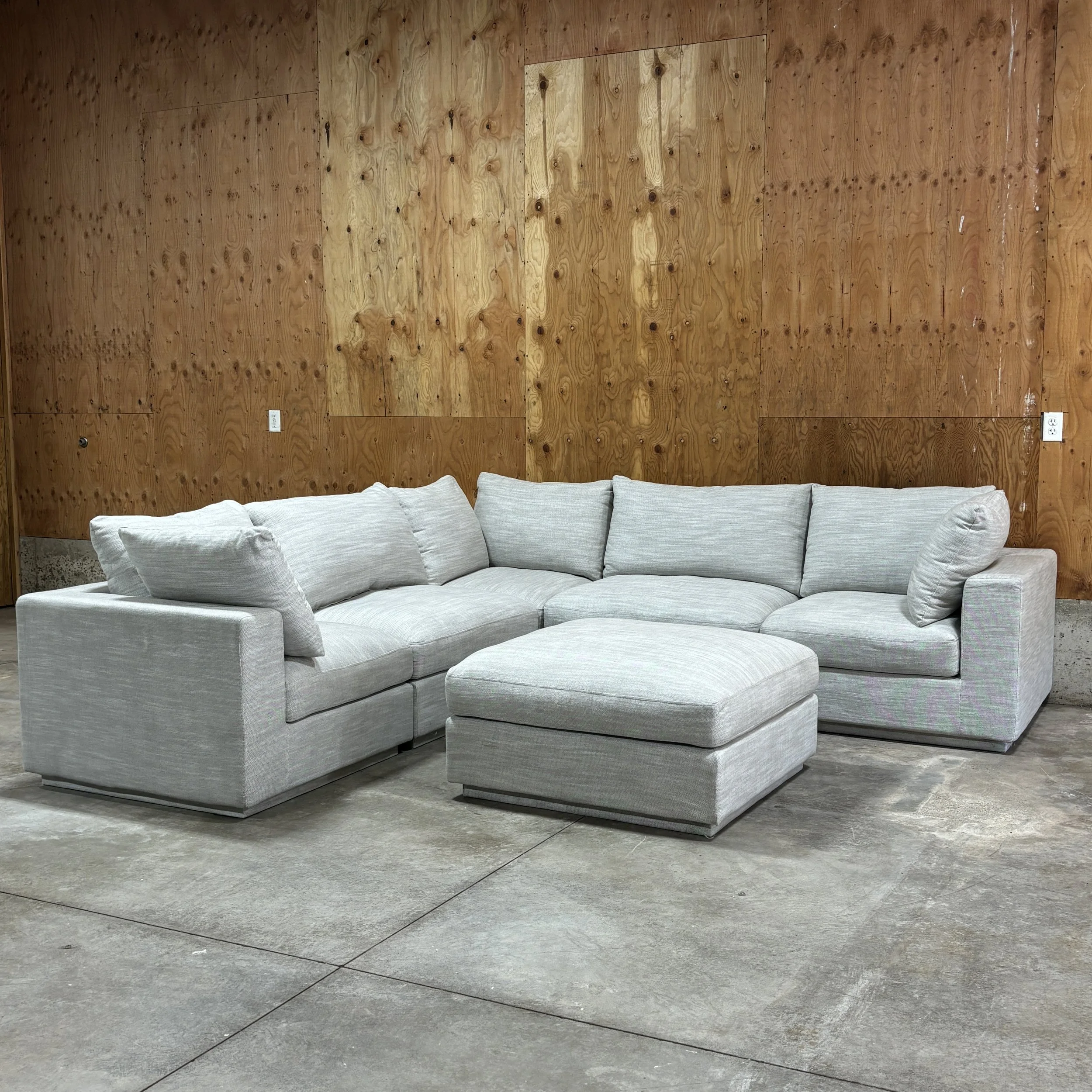 Light gray sectional sofa with ottoman in front, placed against a wooden paneled wall in a room with concrete flooring.