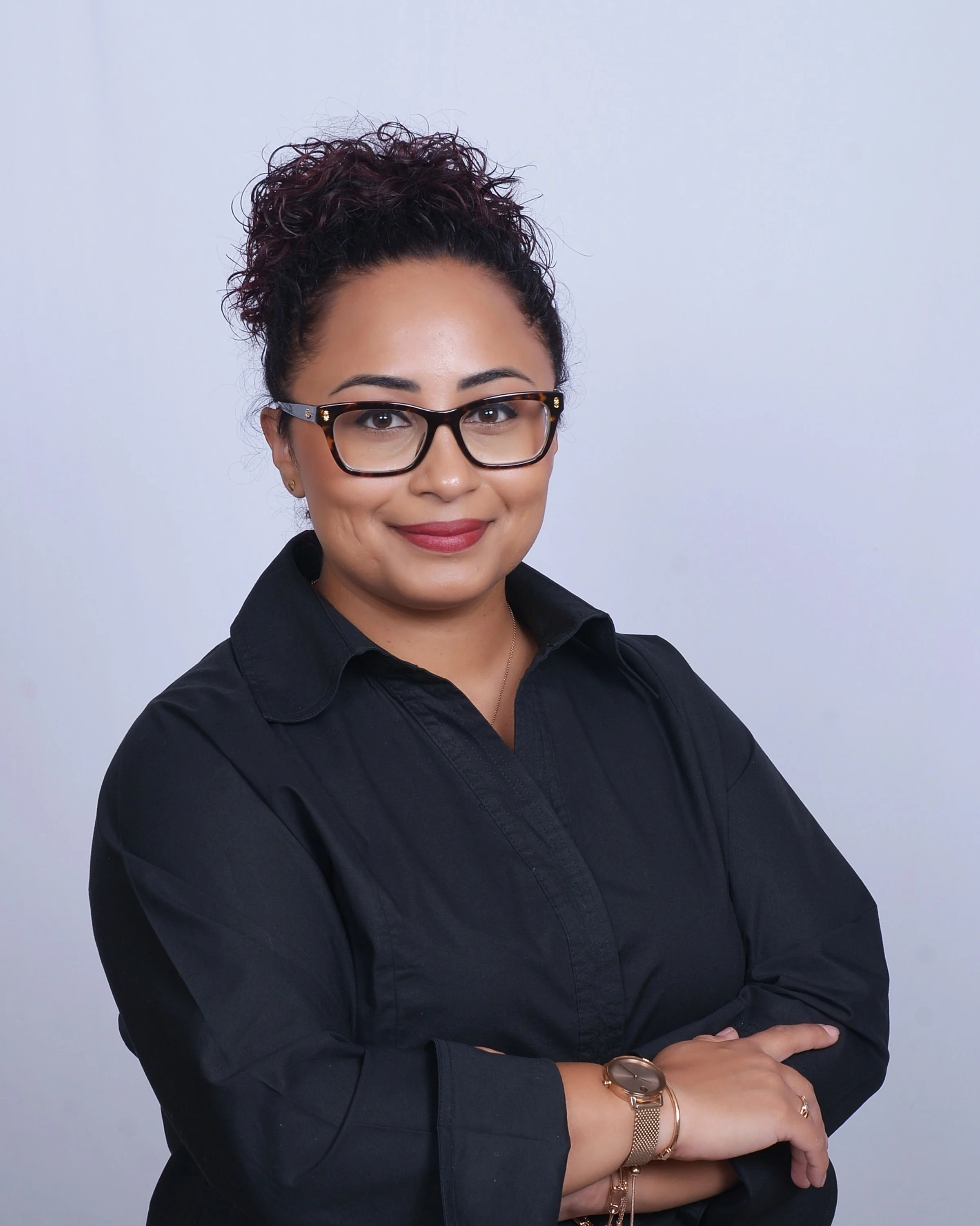 A woman with glasses and curly hair, wearing a black shirt and a watch, is smiling and posing with her arms crossed against a plain gray background.