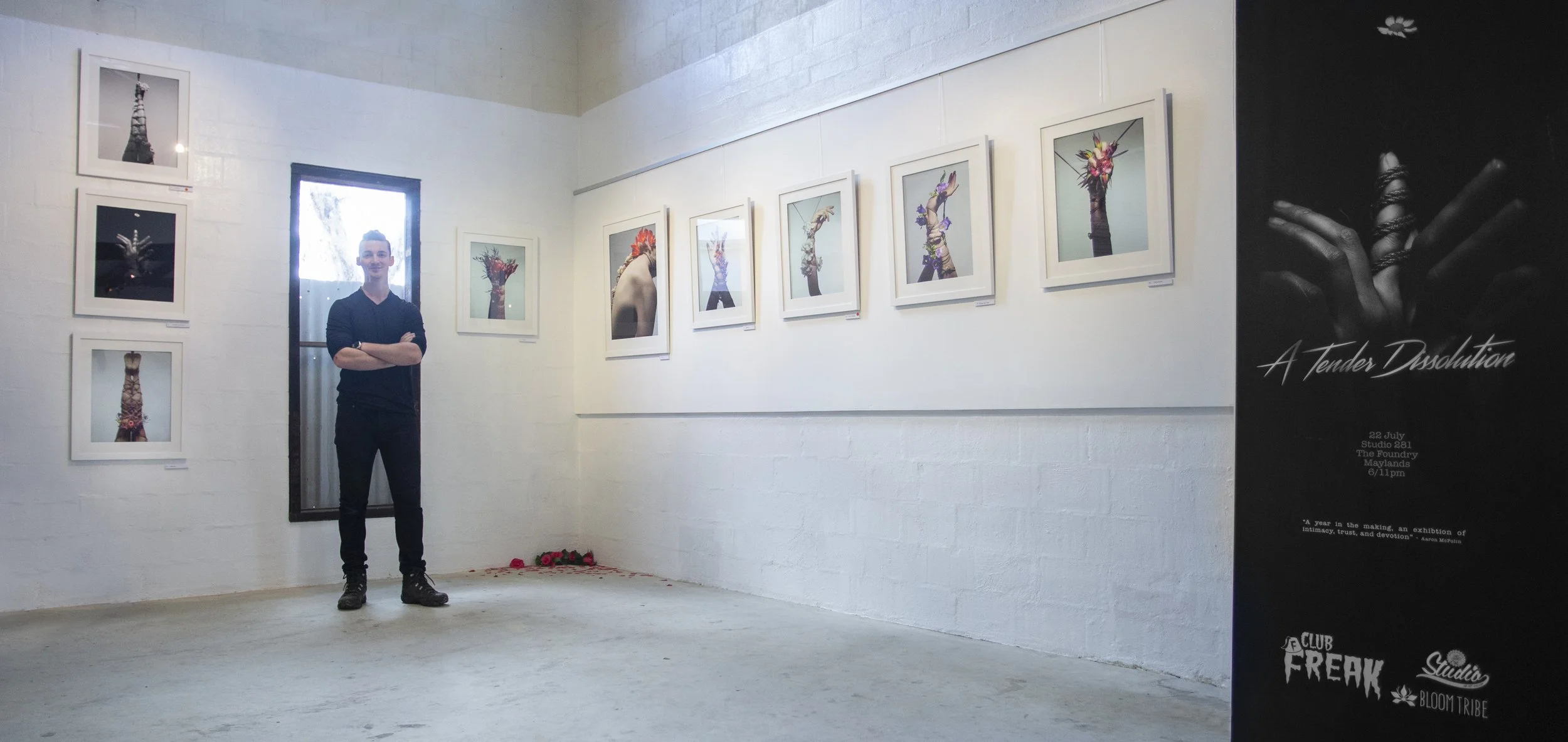 A man standing inside an art gallery next to a wall with framed photographs of hands holding or decorated with flowers, with a poster on the right advertising an art event titled "A Tender Dissolution".