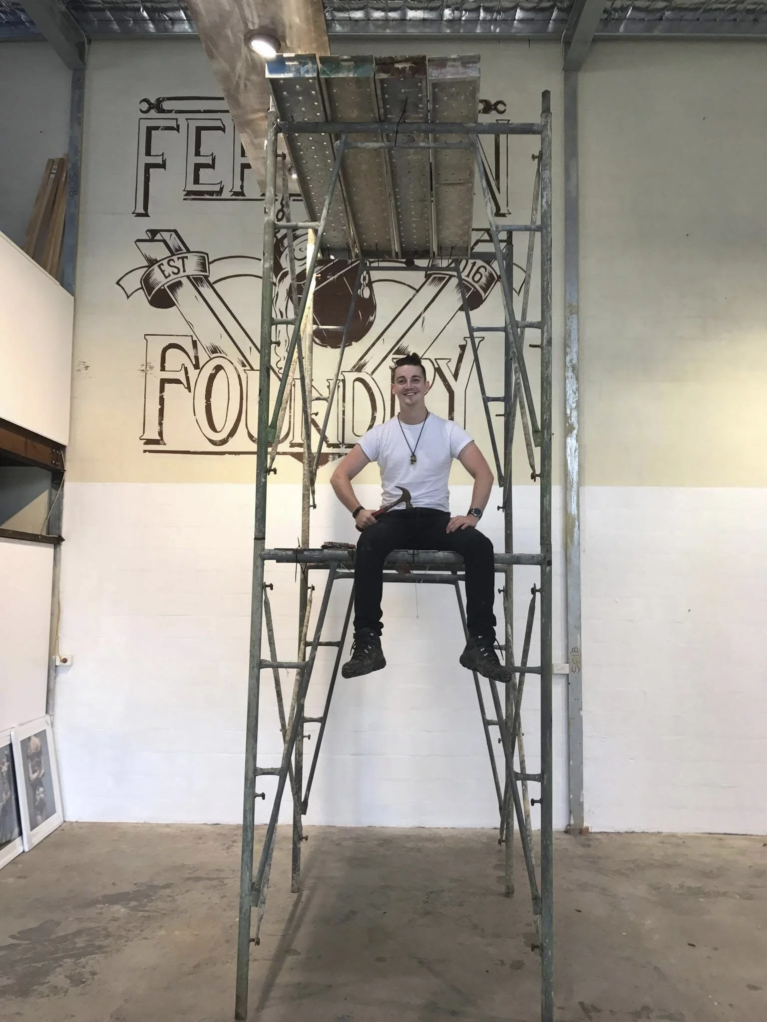 A young man sitting on a metal scaffolding inside a room with unfinished floors and walls. Behind him is a large wall graphic with the text "FOODDY" and decorative elements.