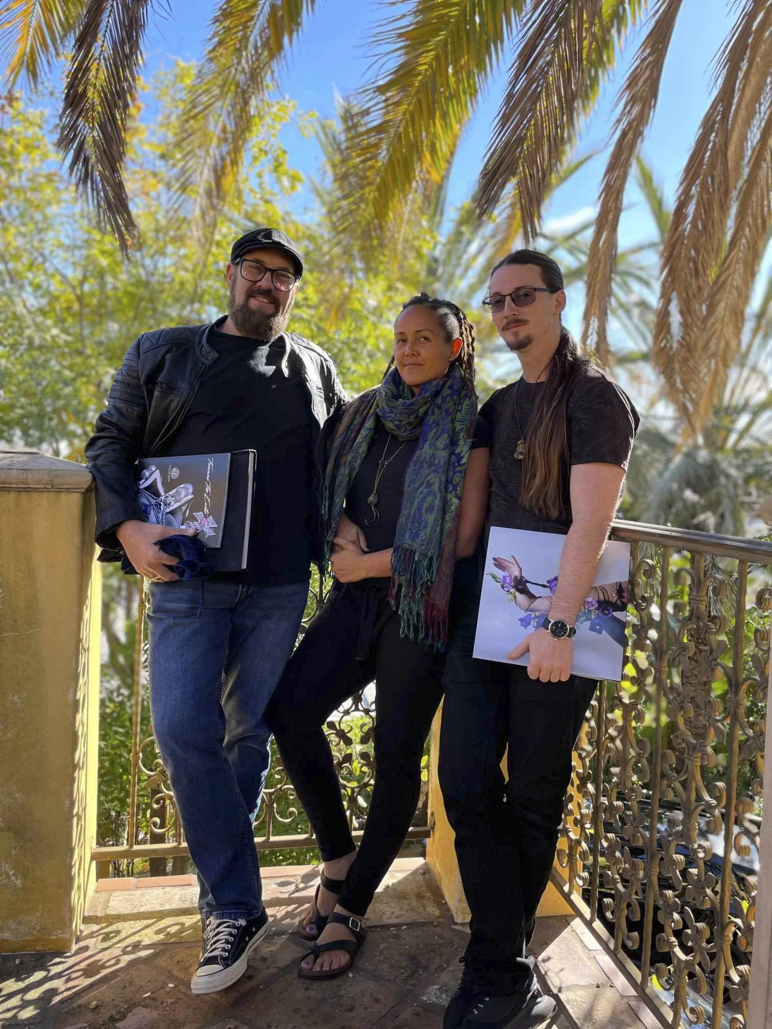 Group of three people standing on a balcony with tropical trees and a blue sky in the background. They are smiling and holding various items, including drawings and a notebook.