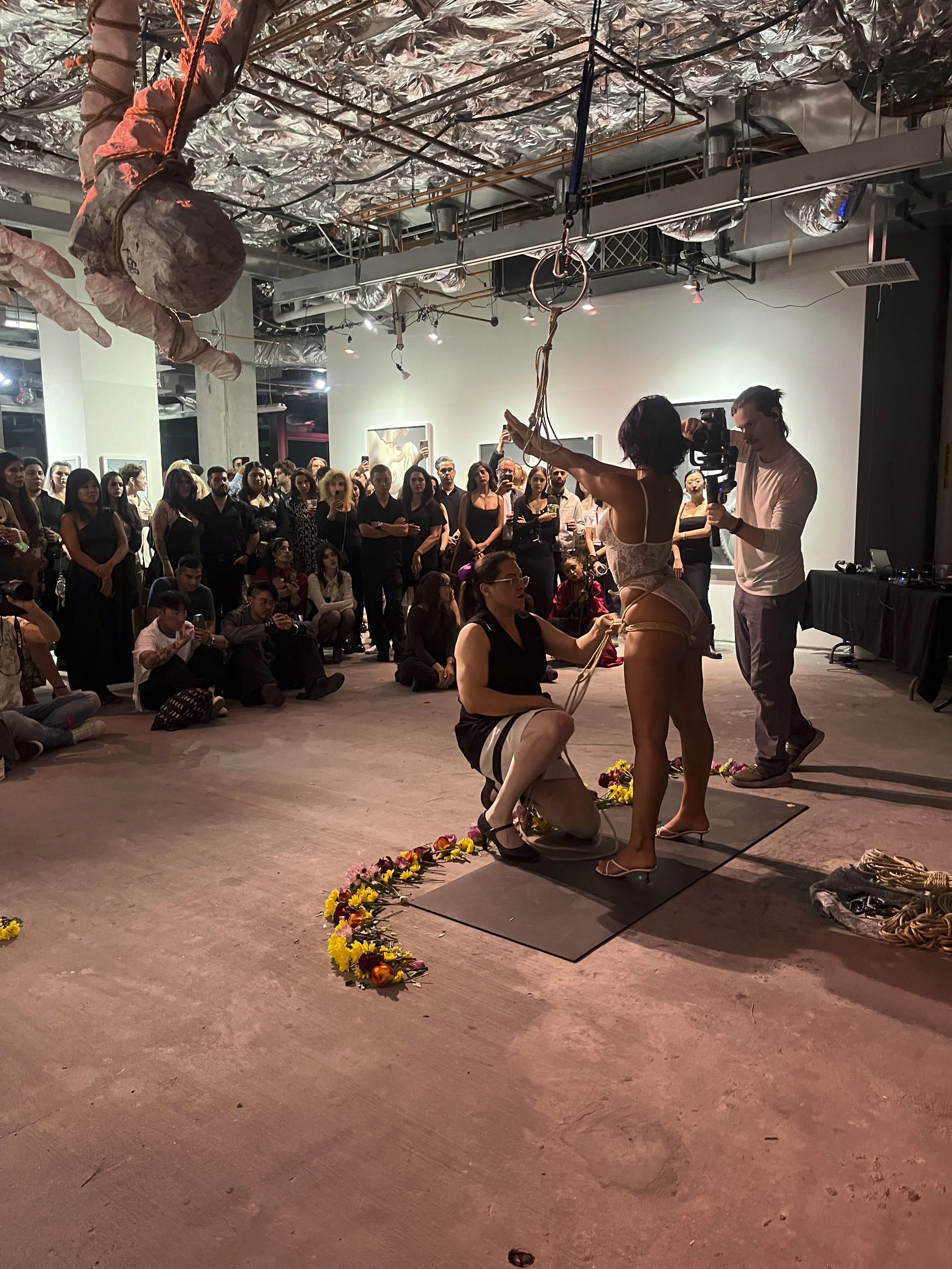 A performance art piece at an art gallery featuring a woman in lingerie holding onto hanging ropes, with another woman kneeling and touching her leg, surrounded by floral arrangements, and a group of diverse spectators observing.