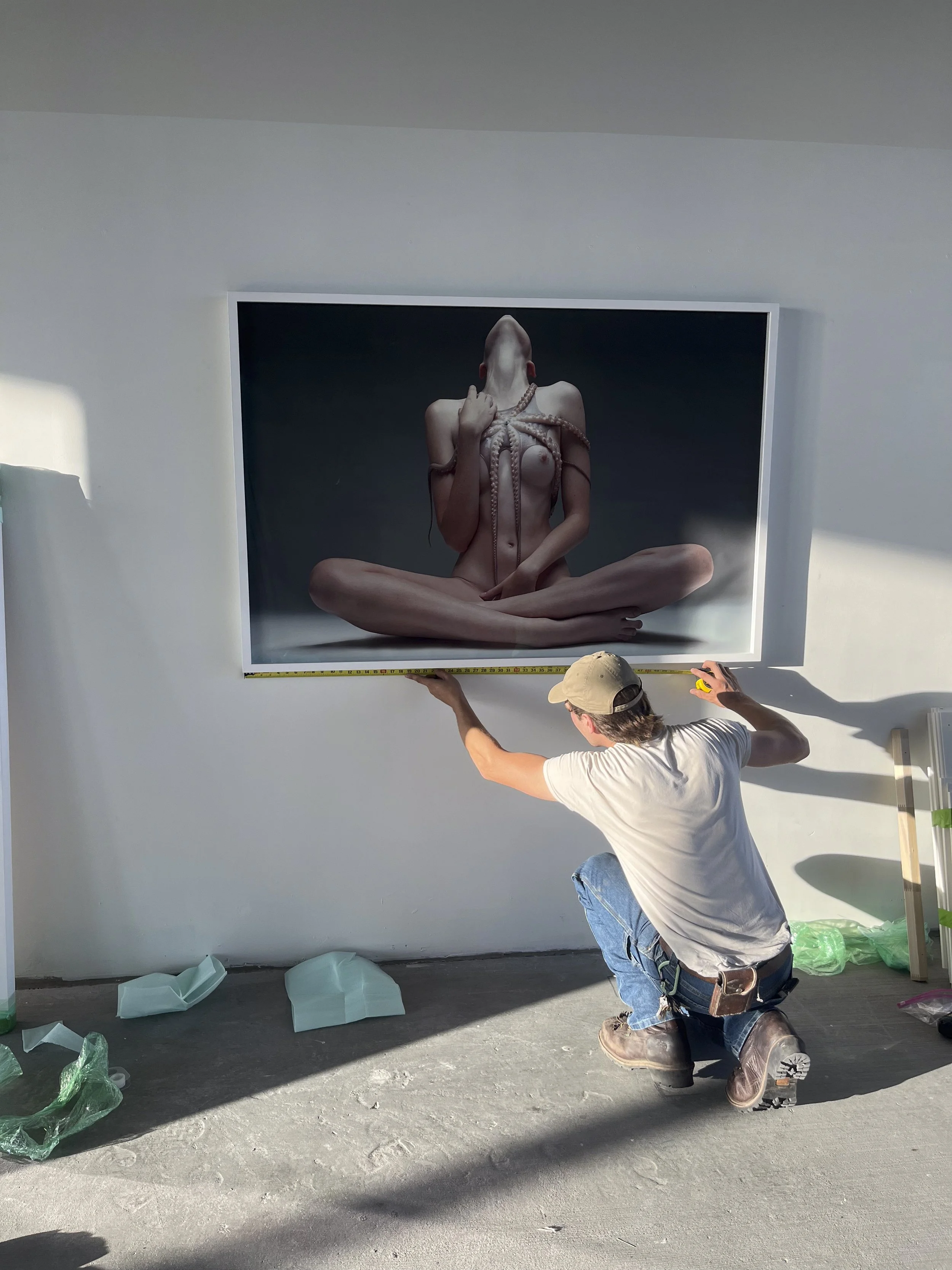 A man installing or adjusting a large framed photo of a woman with a tentacle wrapped around her body on a white wall.