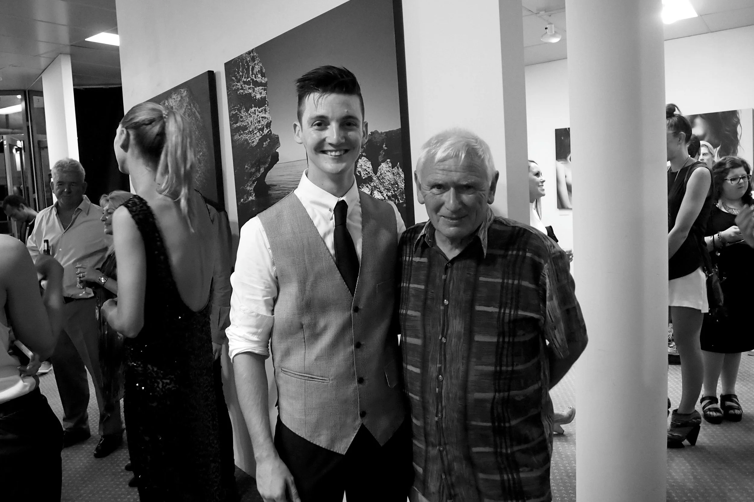 Two men are posing together at an event, with one younger man wearing a vest, tie, and dress shirt and an older man in a patterned shirt. In the background, several other people are talking and enjoying the event, and there are artworks hanging on th