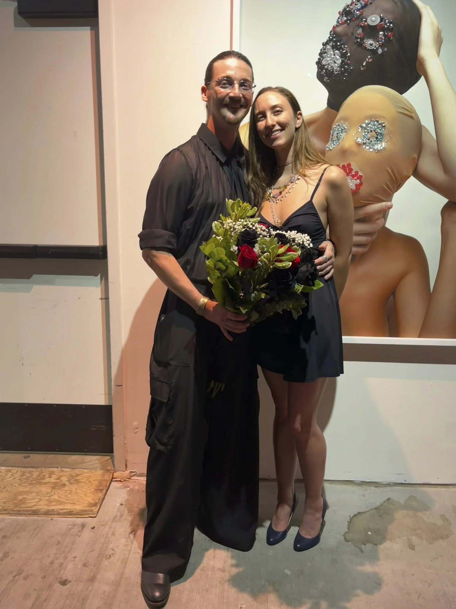 A man and woman are standing together, smiling and holding a bouquet of flowers. The man has long dark hair, glasses, and is wearing a black shirt and pants. The woman has long brown hair and is dressed in a black dress with thin straps and high heel