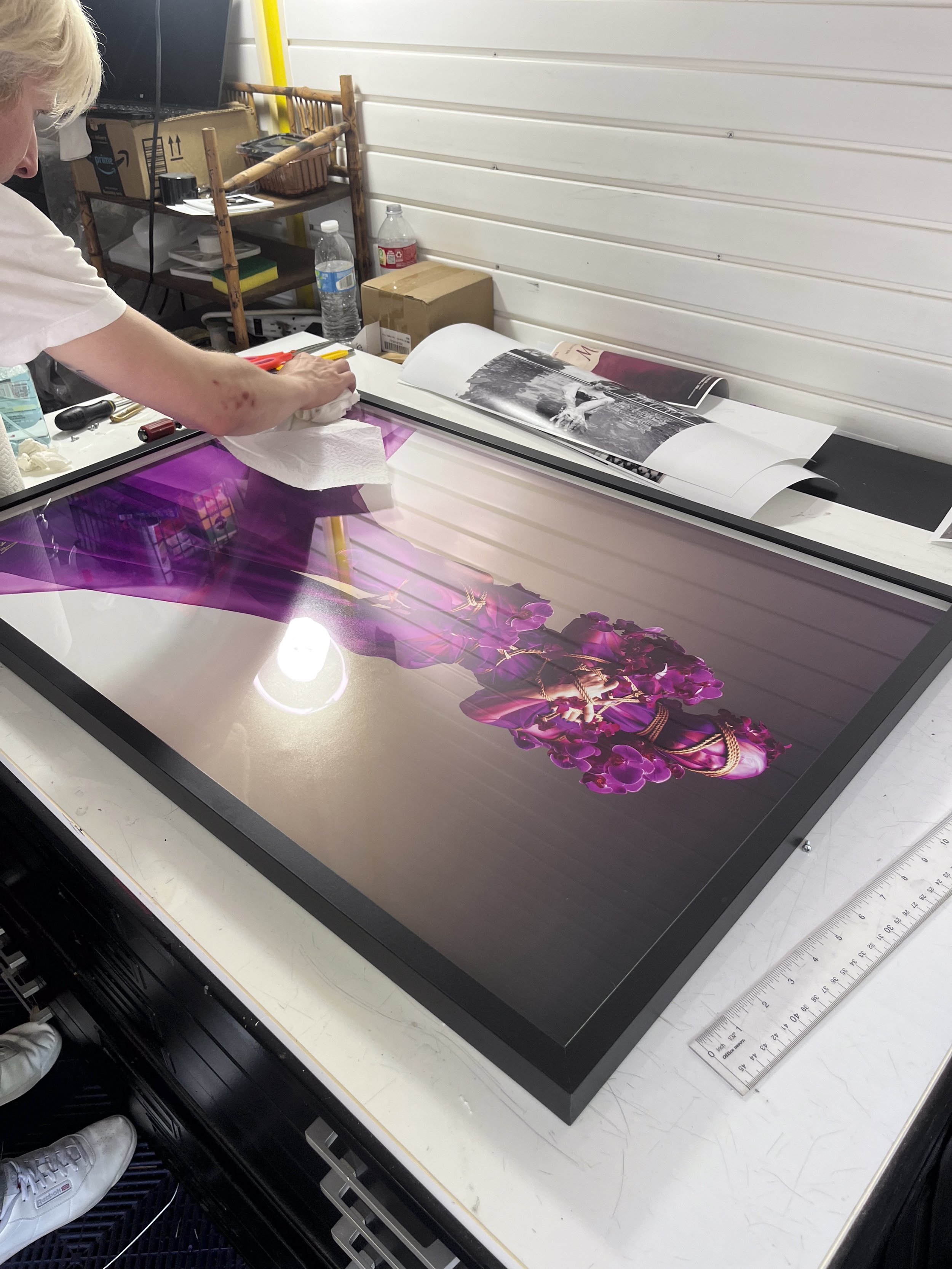 Person cleaning or inspecting a large, vibrant, purple-flowered artwork or print on a flat surface in a workspace.