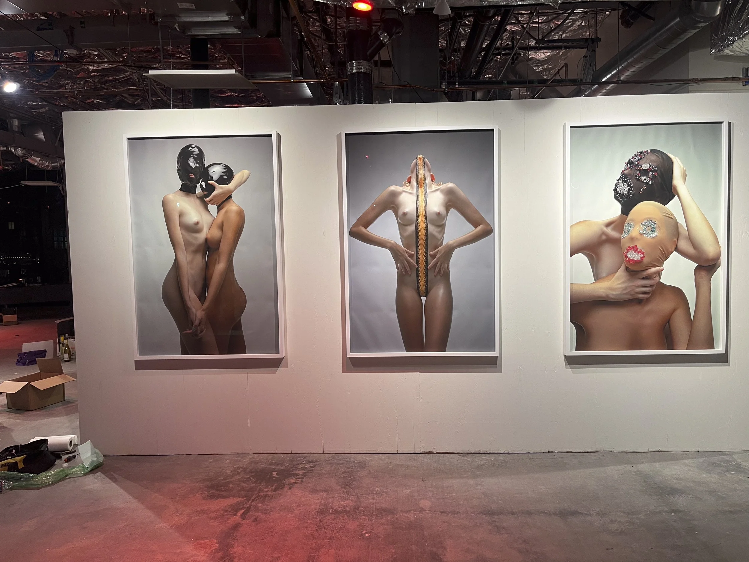 Three framed photographs of partially nude models with masks or masks decorated with various ornaments on display on a white gallery wall.