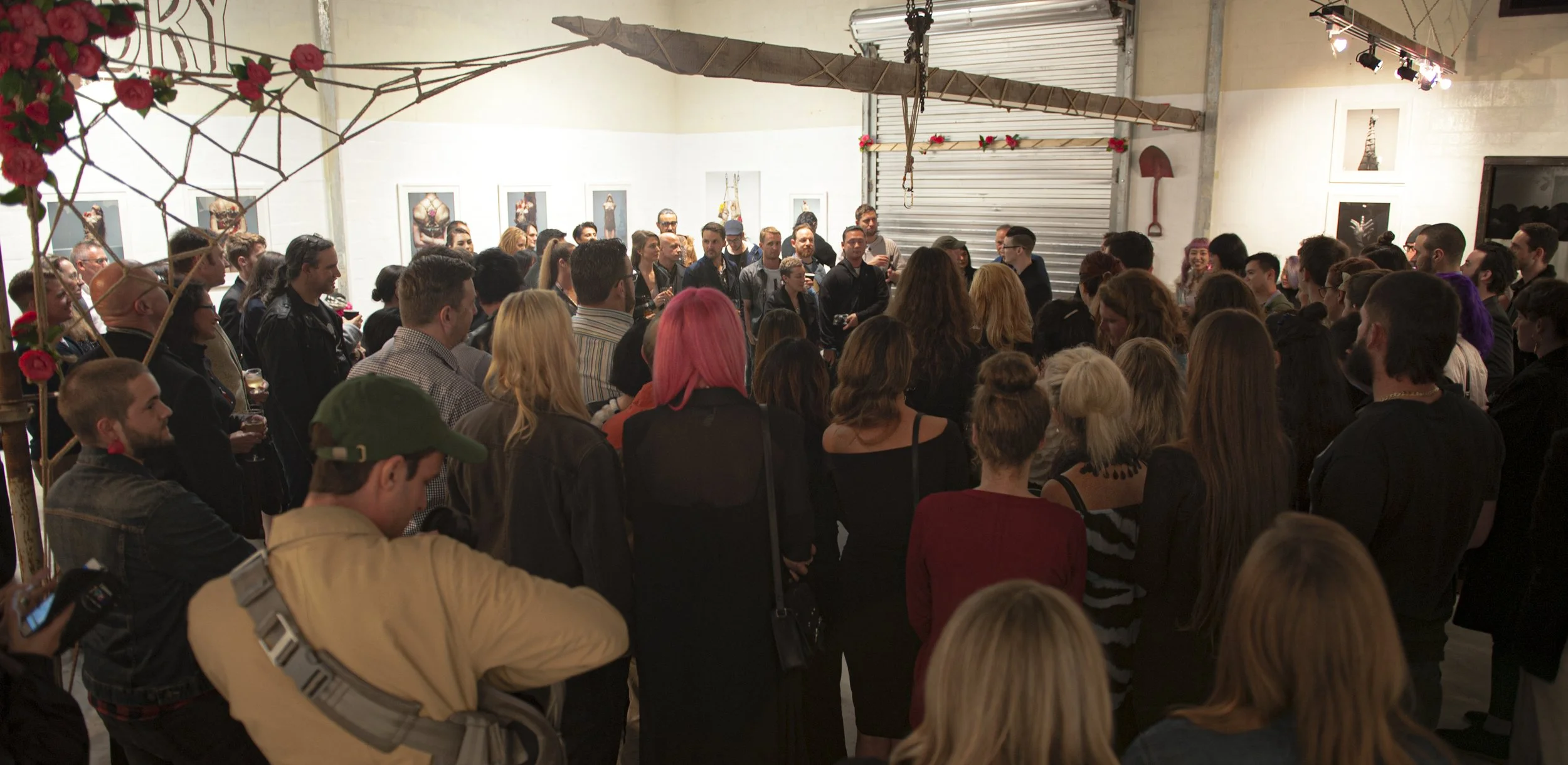Crowd of people attending an art exhibition or event in a gallery with framed artwork on the walls.