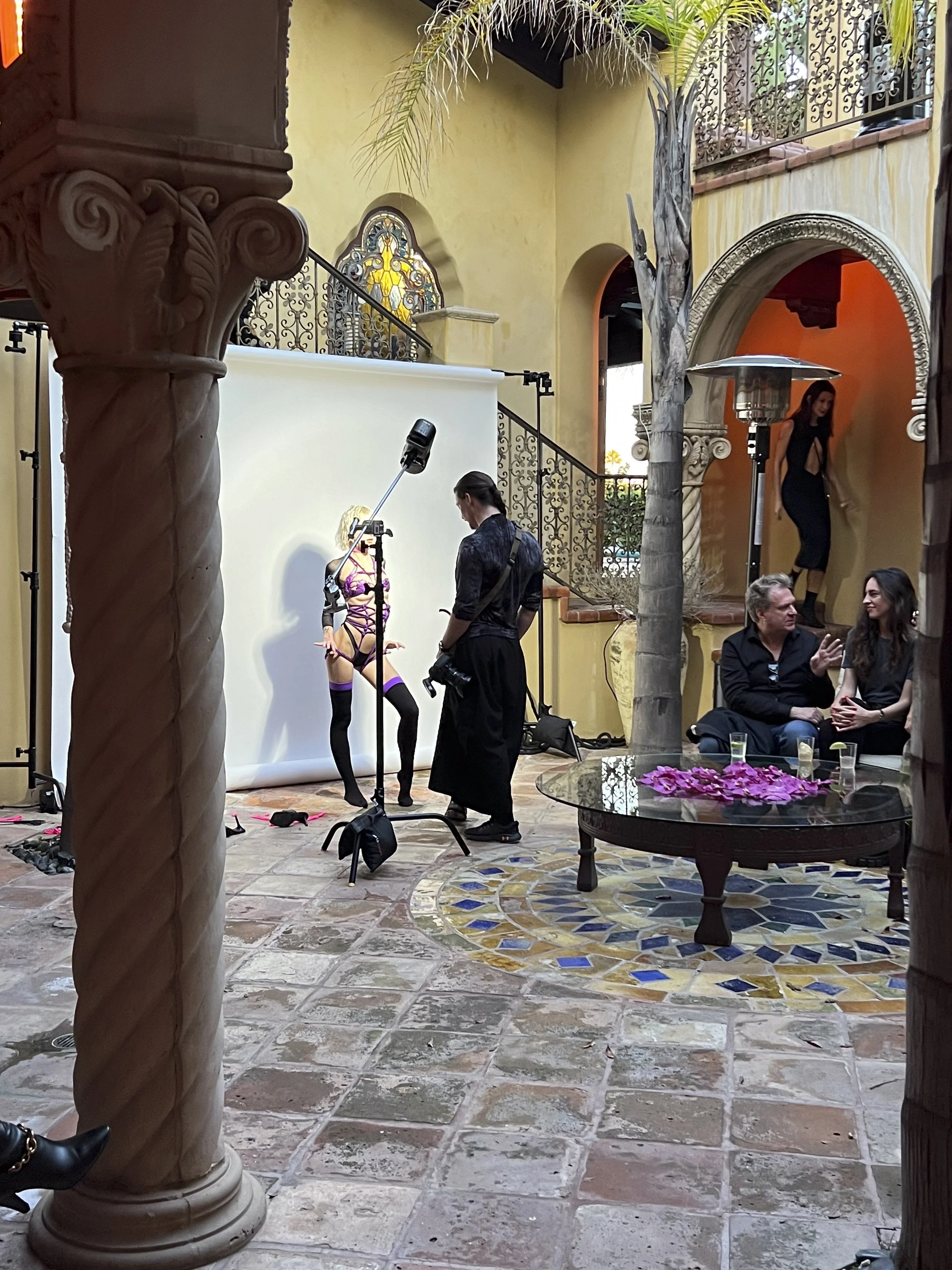 Photoshoot scene in a courtyard featuring a woman in lingerie, photographers, and onlookers, with decorative architectural elements and stained glass windows.