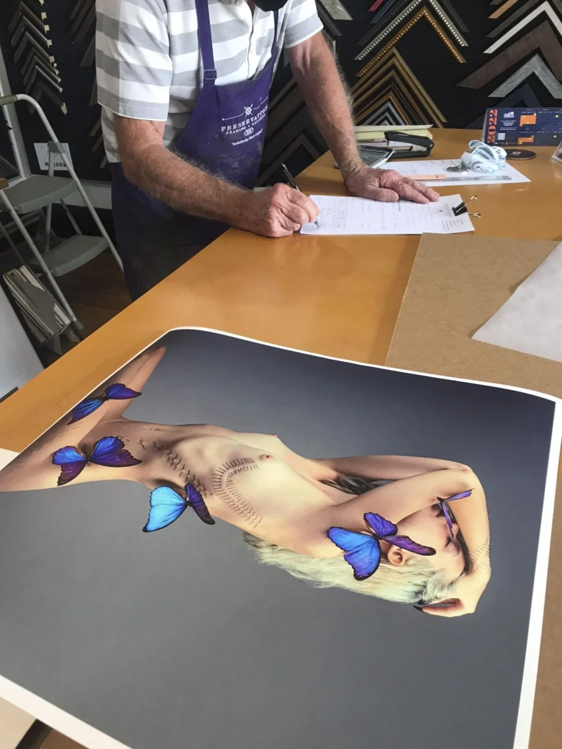 A person's hands and upper body are visible as they sign documents on a wooden table. In the foreground, there is a large print of a digital artwork of a nude woman with blonde hair, covered with blue and purple butterflies.