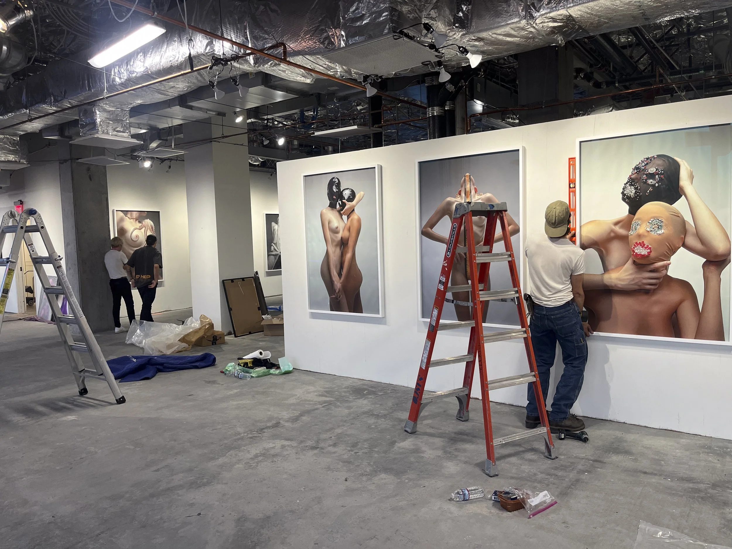 Gallery workers setting up an art exhibition with large photographs of women wearing masks and covering their bodies. The space is industrial with exposed ducts and scaffolding, and various tools and supplies are scattered on the floor.